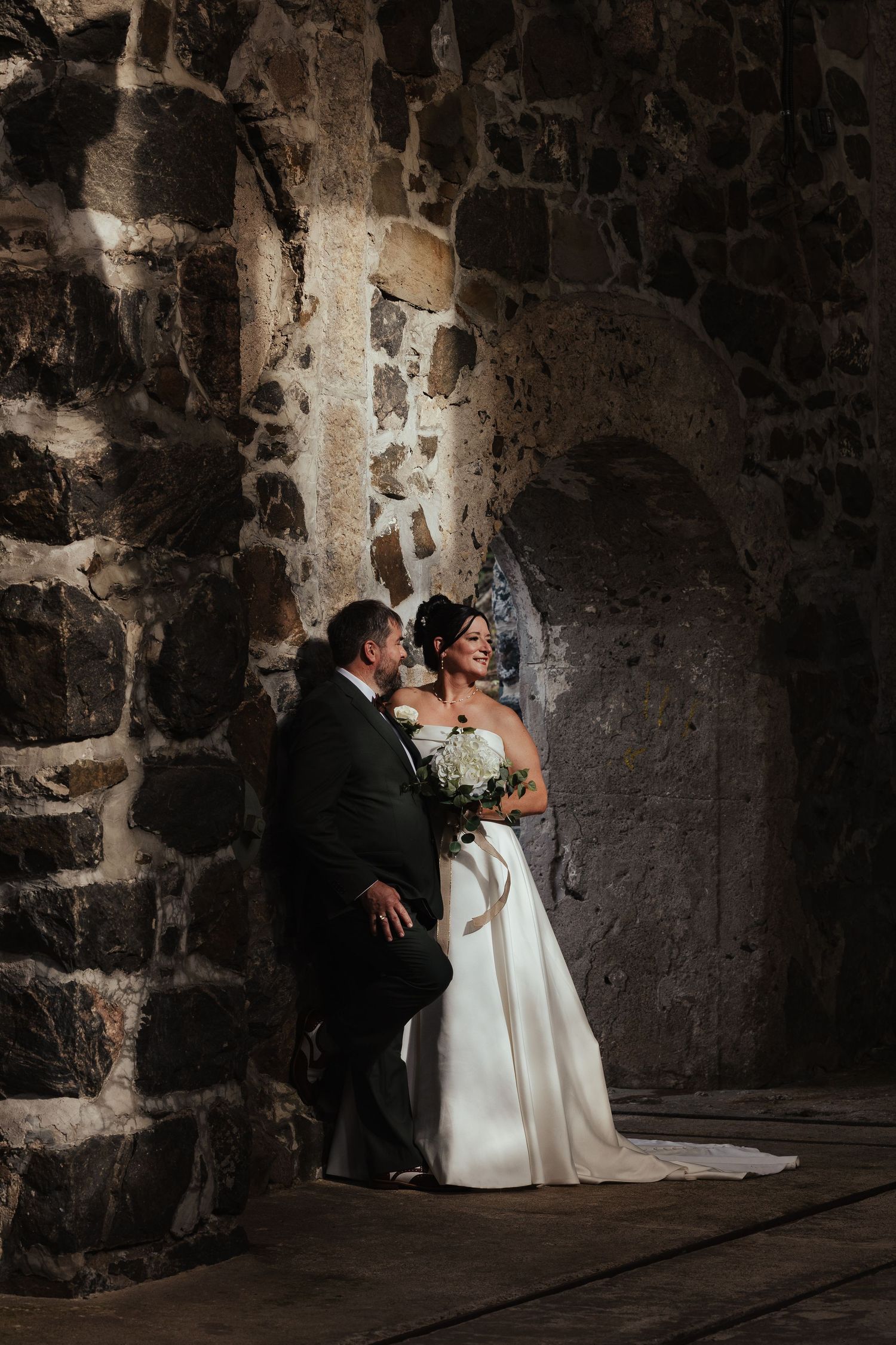 Bride and groom share romantic moment leaning against rustic stone wall in dramatic lighting during wedding portrait.
