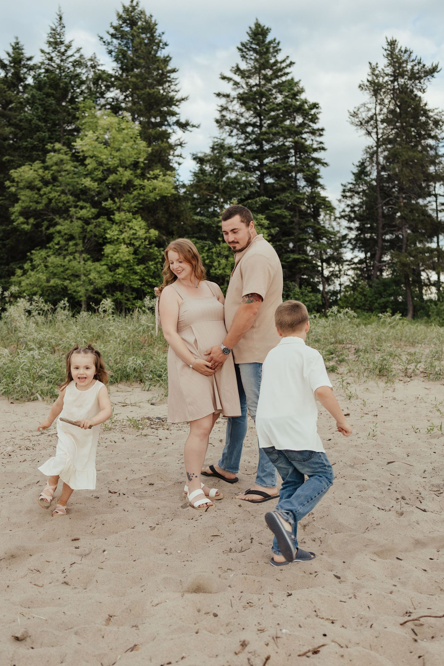 Pregnant woman and man hold hands on sandy beach while two young children play around them with pine trees in background.