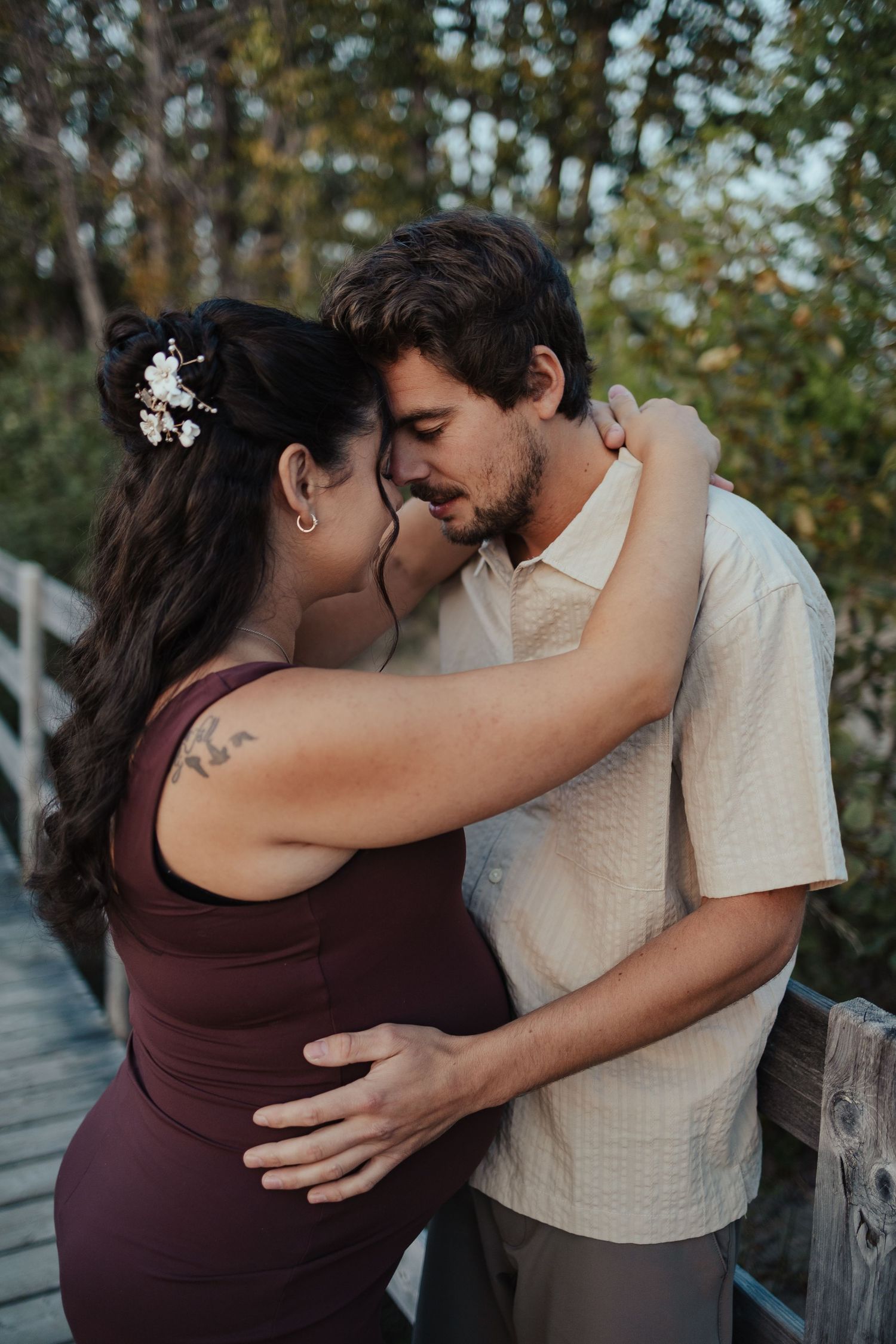 Couple sharing a tender kiss on a wooden bridge, woman in burgundy dress with floral hair accessory, man in beige shirt.