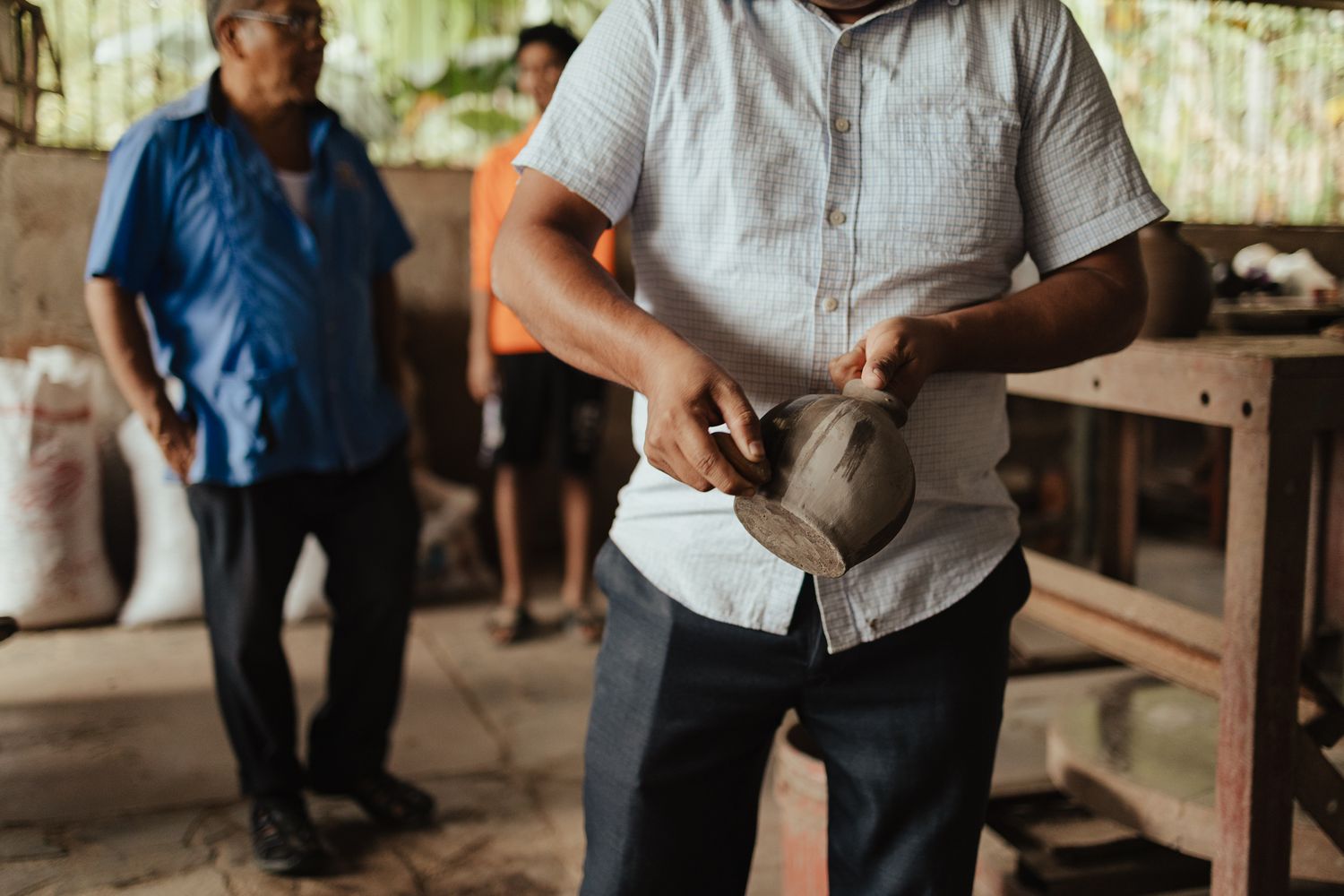 Man in white shirt holds a clay pot in a rustic workshop while another man stands in the background.