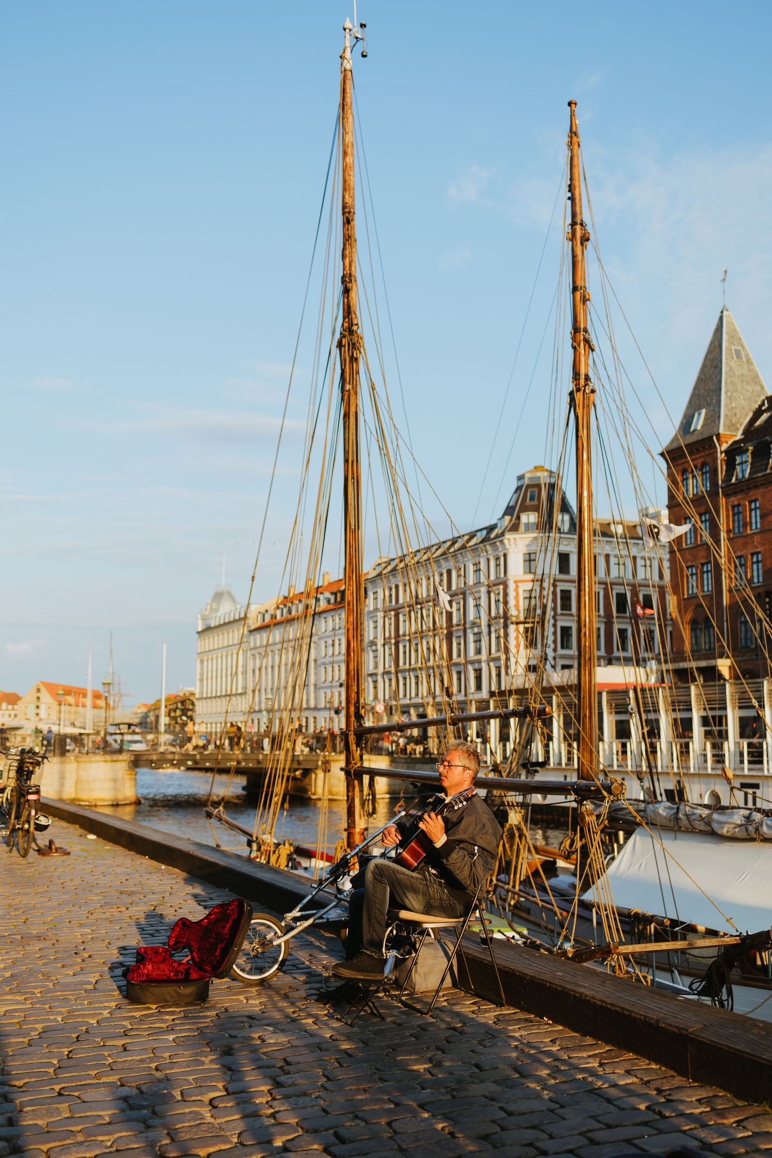 Person sitting on cobblestone dock beside tall sailing ships in a historic European harbor at golden hour.