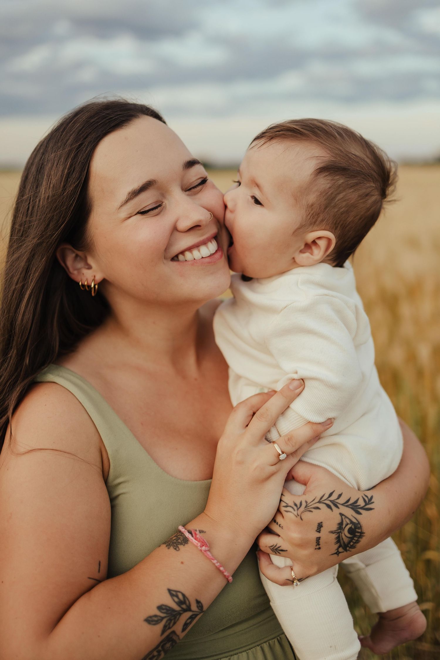 A smiling young mother with tattoos holds her baby in a golden field as the infant gives her a sweet kiss.