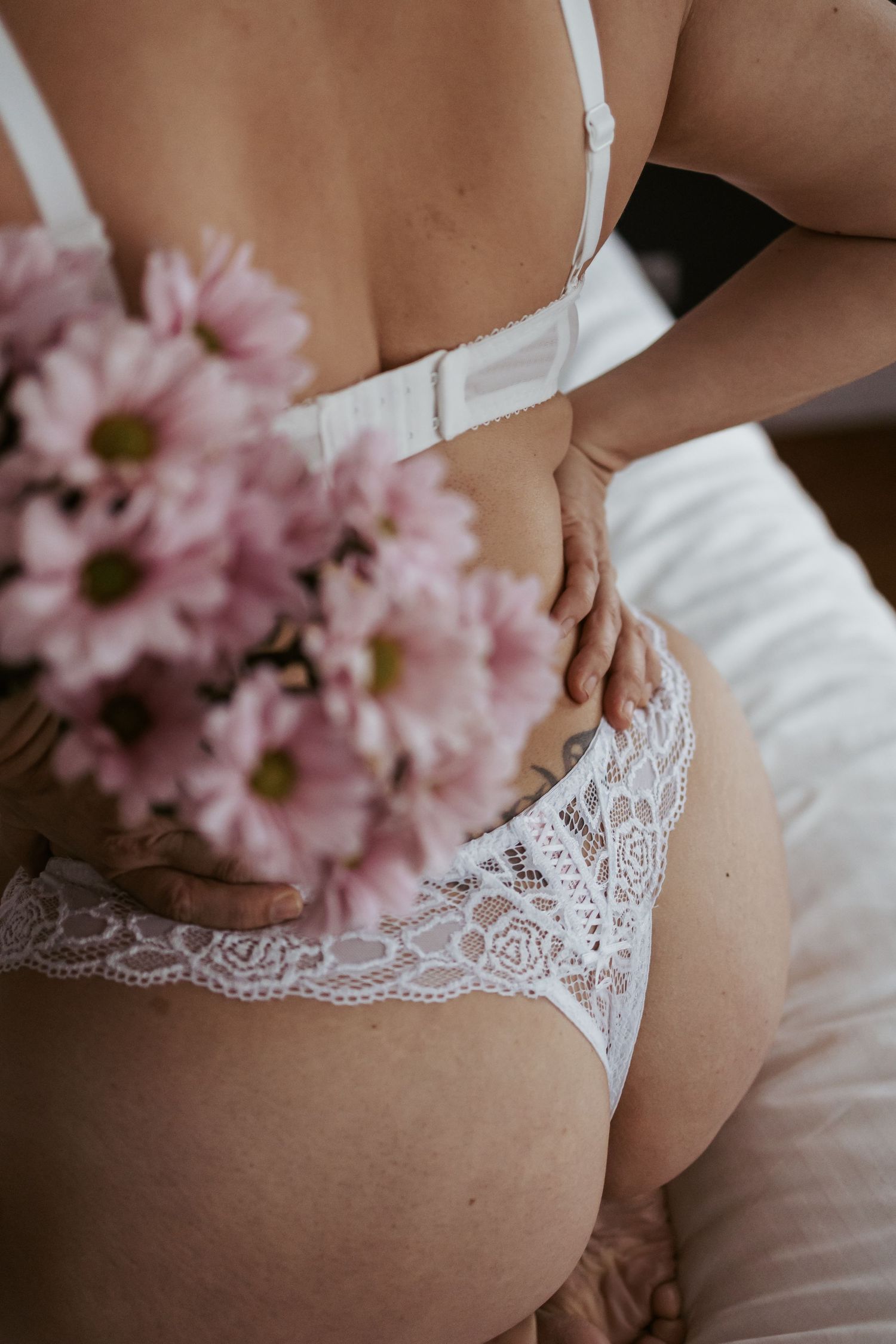 Woman in white lace lingerie holding pink chrysanthemum flowers, boudoir photography on white bedding.