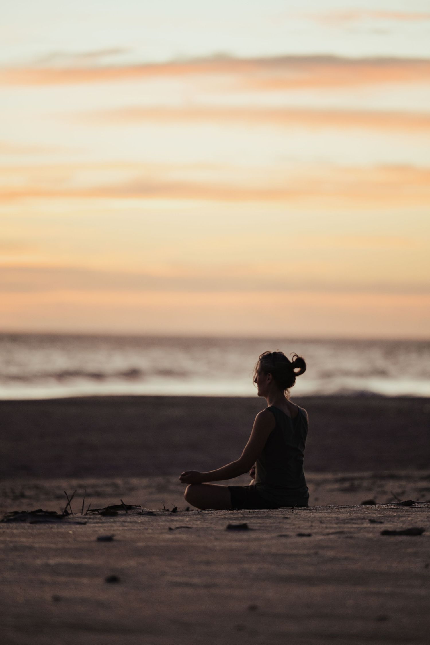 Woman meditating in lotus pose on a sandy beach at sunset, silhouetted against a colorful orange sky.