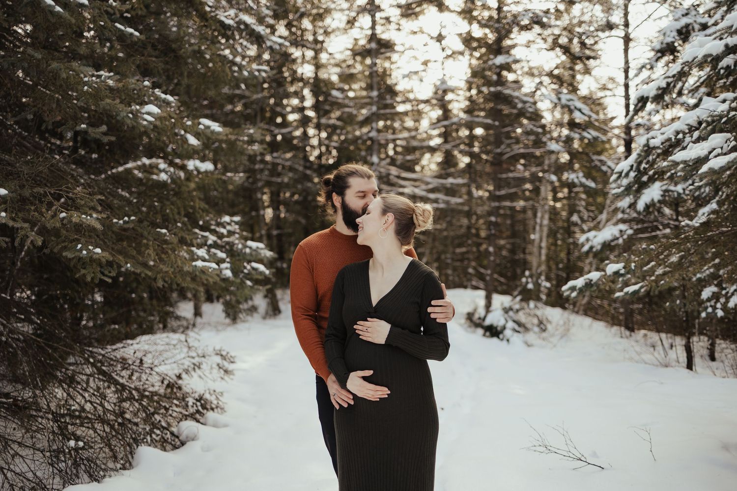 Couple embracing in snowy winter forest, pregnant woman in black dress with partner in red sweater.