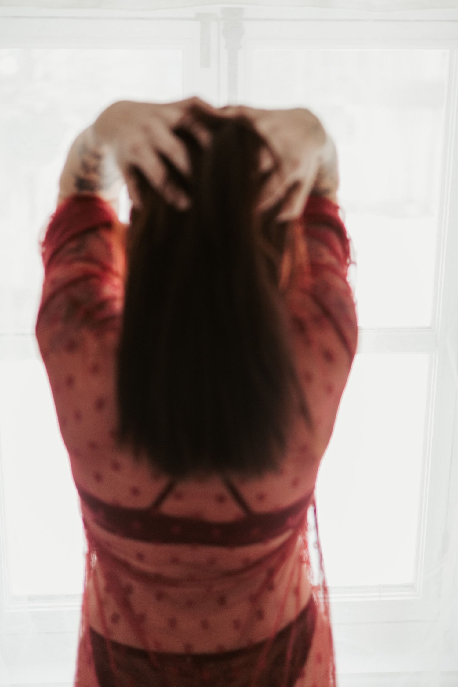 Woman in red lace dress standing at window, hands in dark hair, viewed from behind in soft focus.