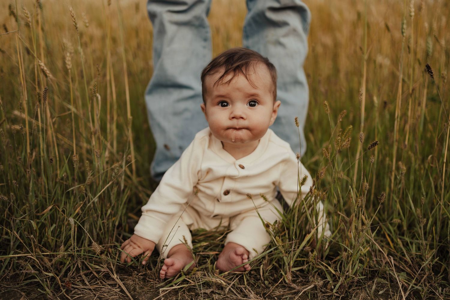 Adorable baby sitting in tall grass field wearing cream romper, looking up curiously with adult standing behind.