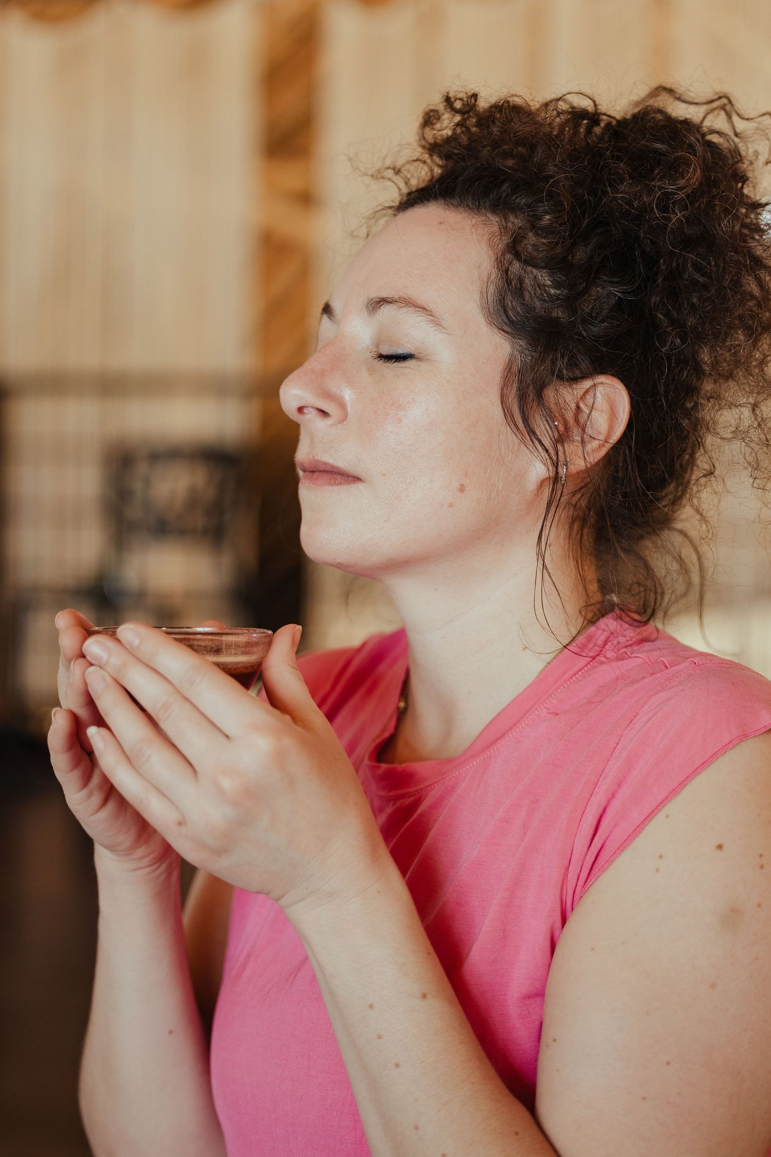 Woman in pink top holding a glass cup with eyes closed, savoring the aroma of a warm drink indoors.
