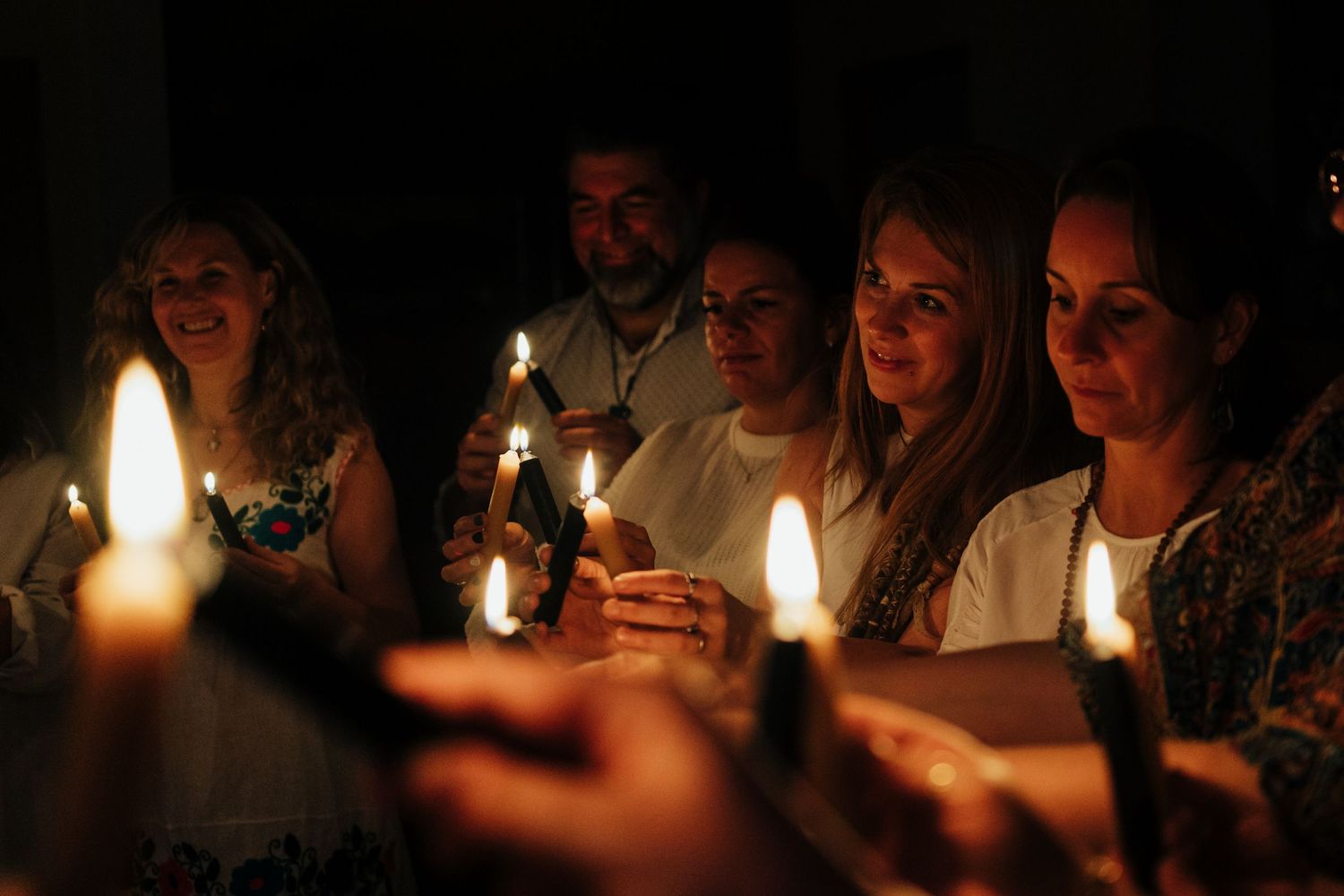 Group of smiling people holding lit candles together in a dark room during a candlelight ceremony.