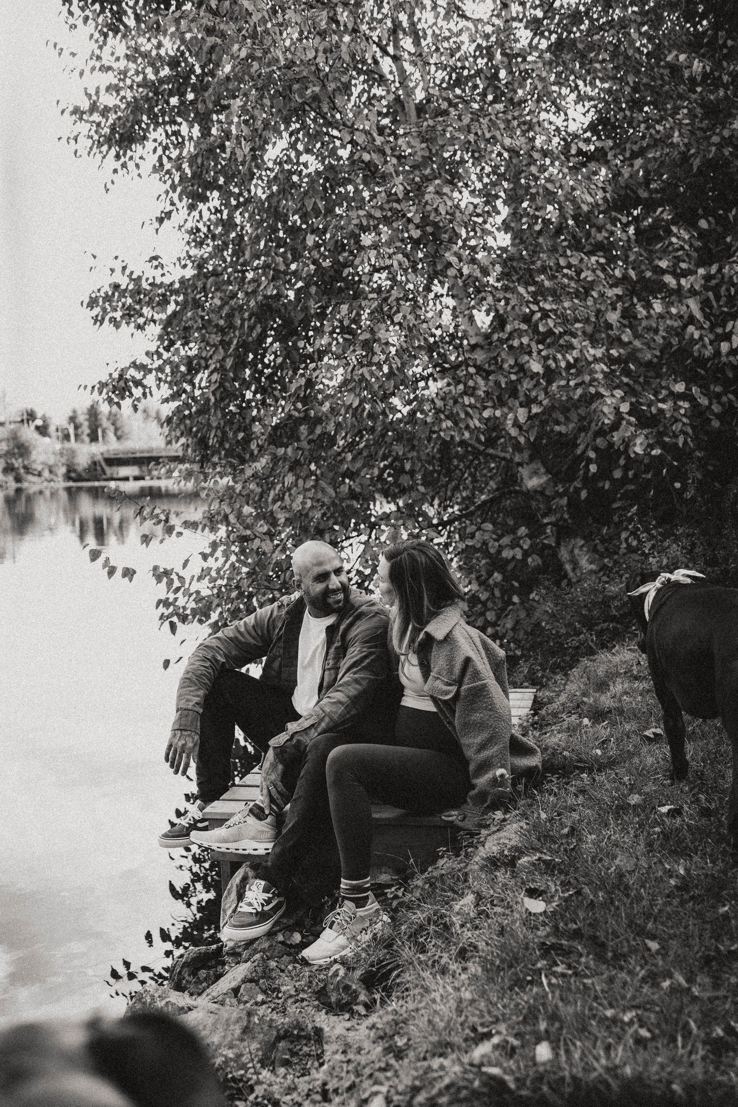 Black and white photo of a couple sitting together by a lakeside under trees, sharing an intimate moment outdoors.