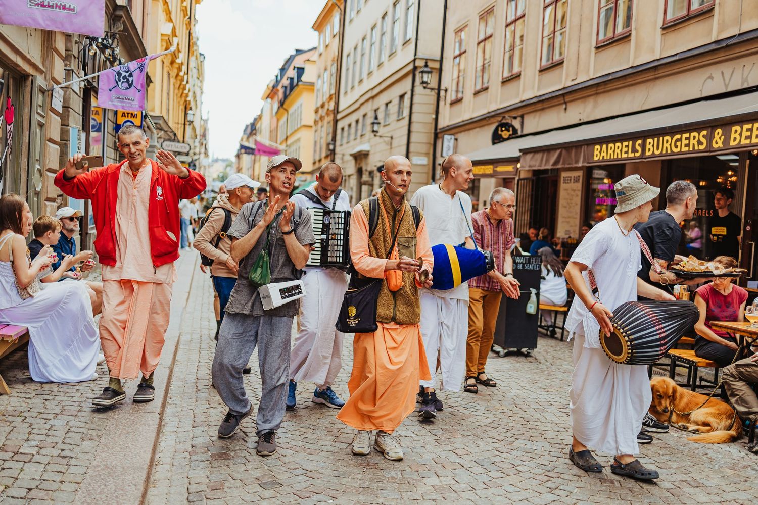 Hare Krishna devotees chanting and playing instruments on a busy European pedestrian street lined with shops.