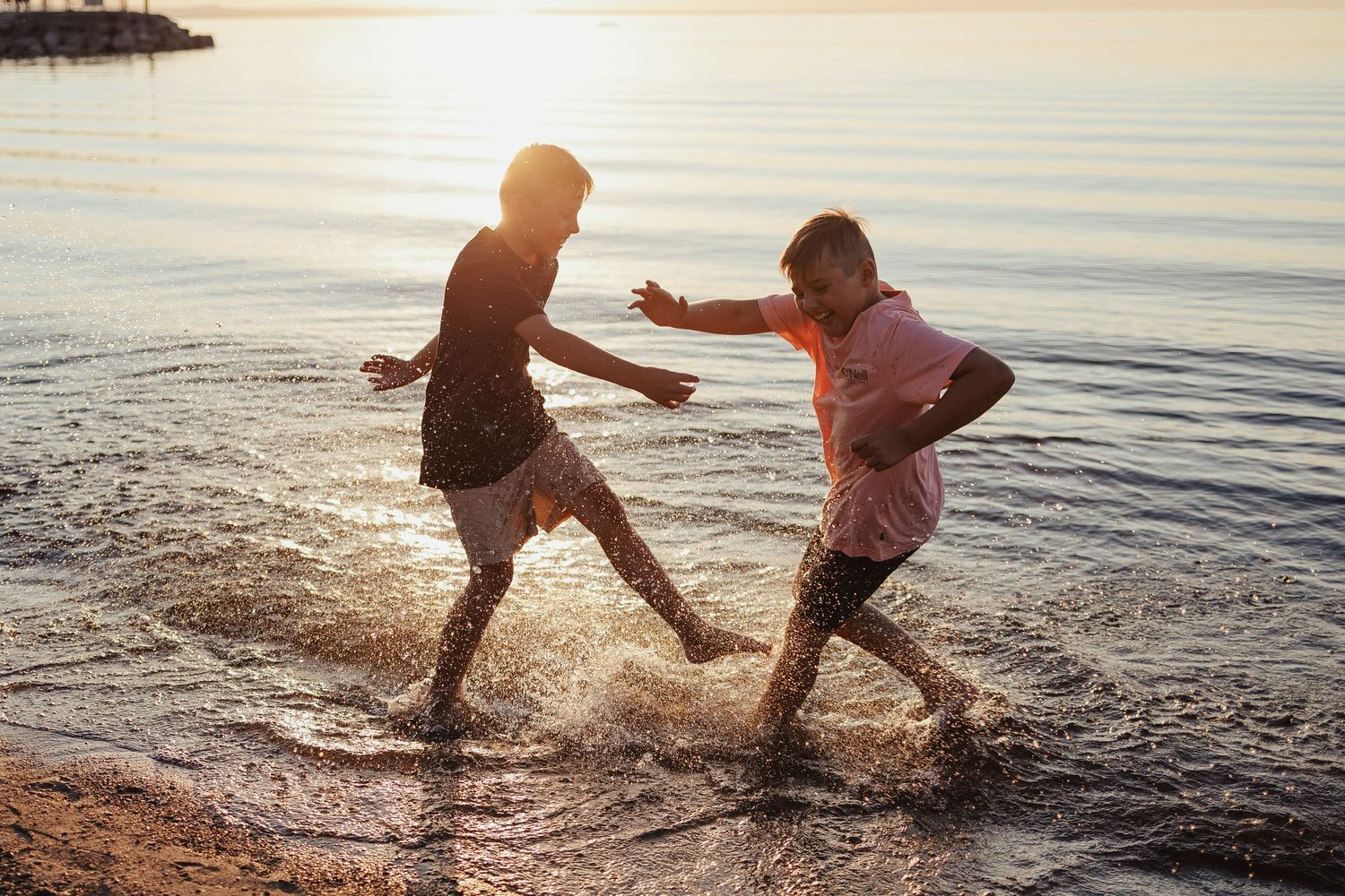 Two children splashing and playing in shallow ocean waves at sunset on a sandy beach.