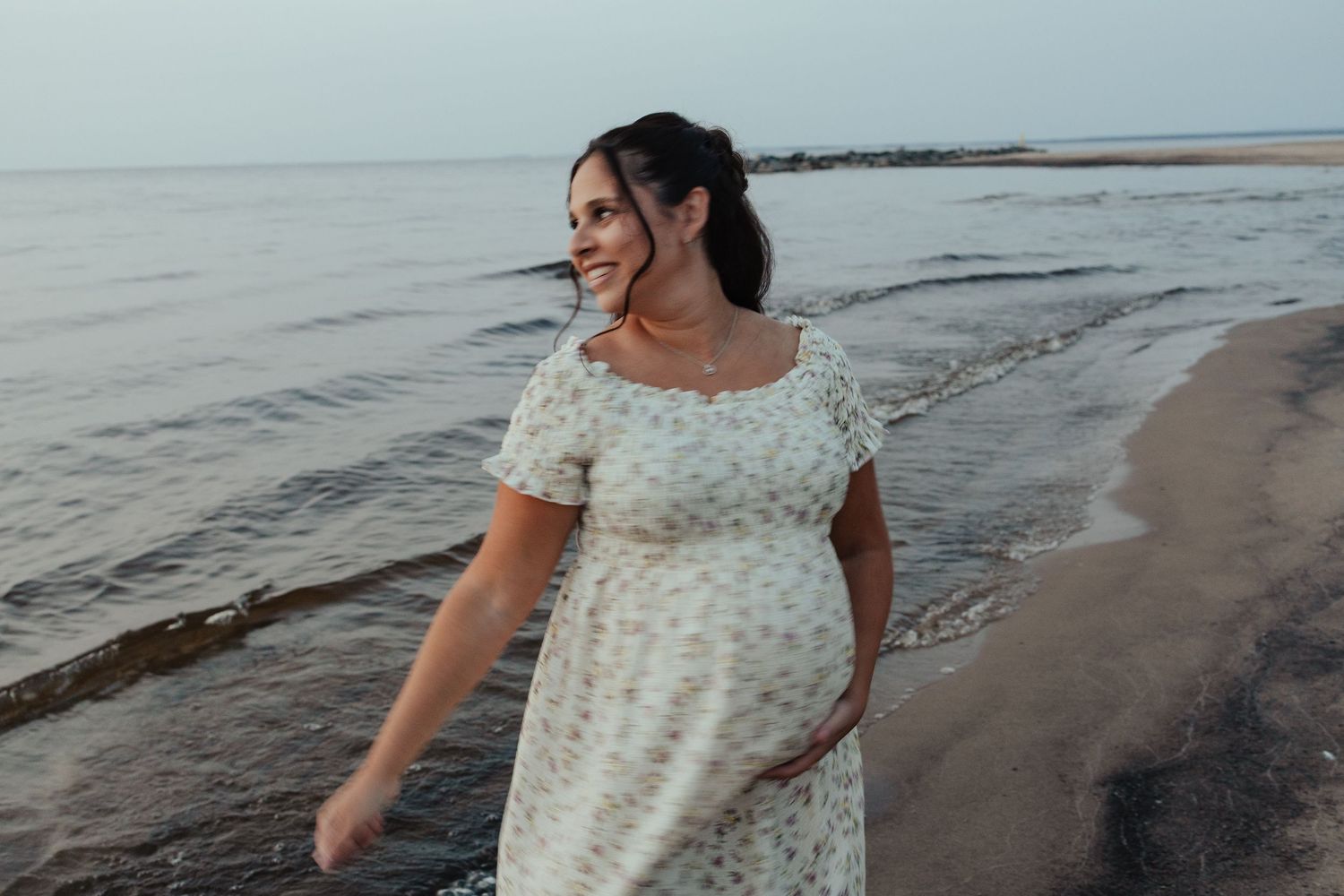 Smiling pregnant woman in floral dress walking along a serene beach shoreline at dusk.