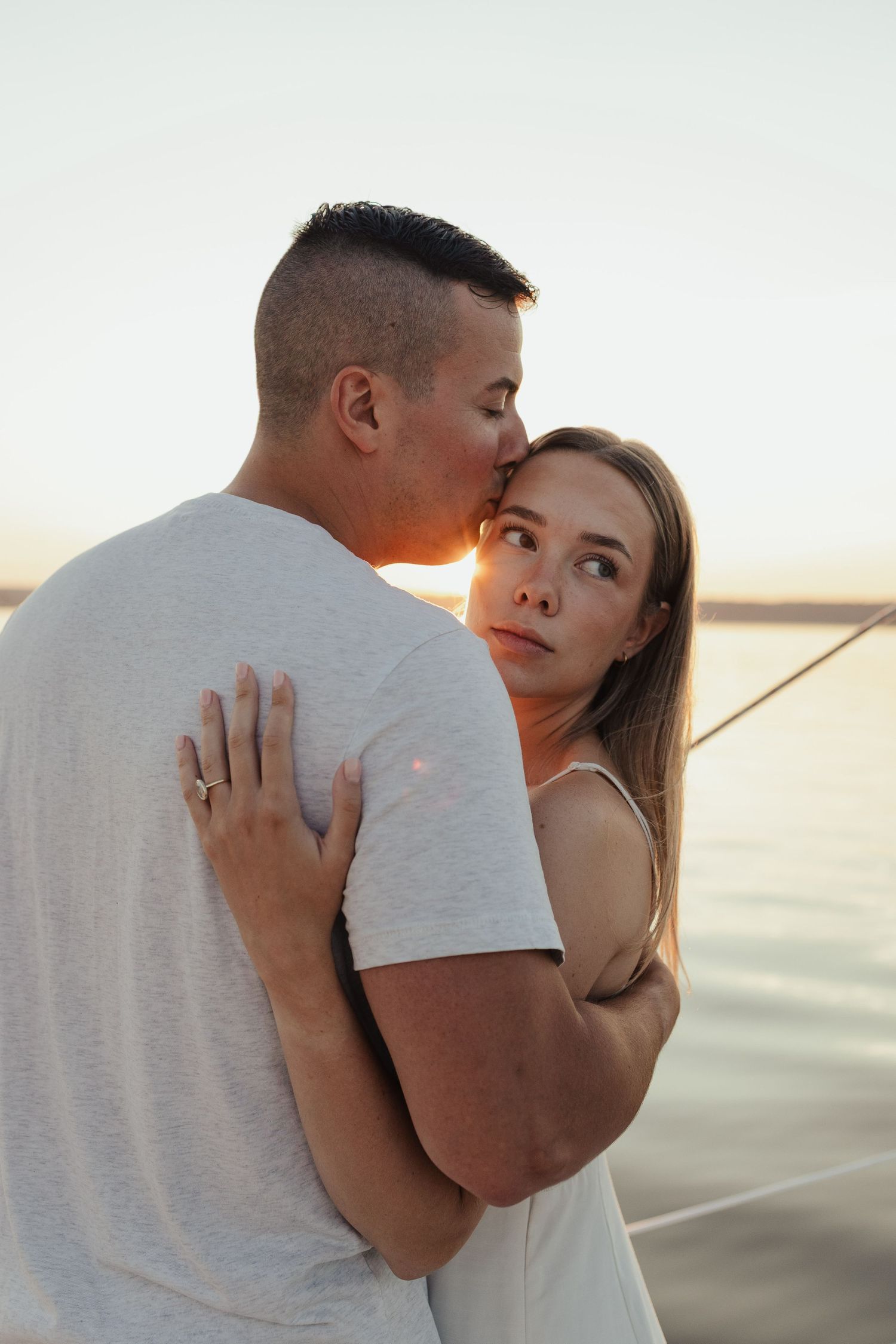 Couple embracing at sunset by the water, man kissing woman on the cheek, romantic golden hour portrait.