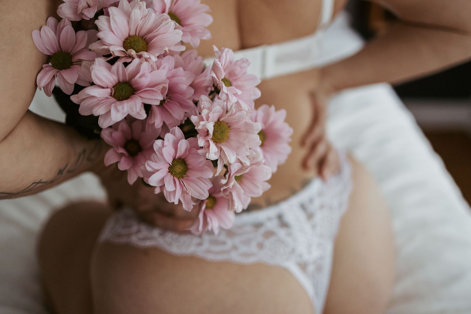 Person in white lace lingerie holding pink daisy flowers while sitting on a bed.