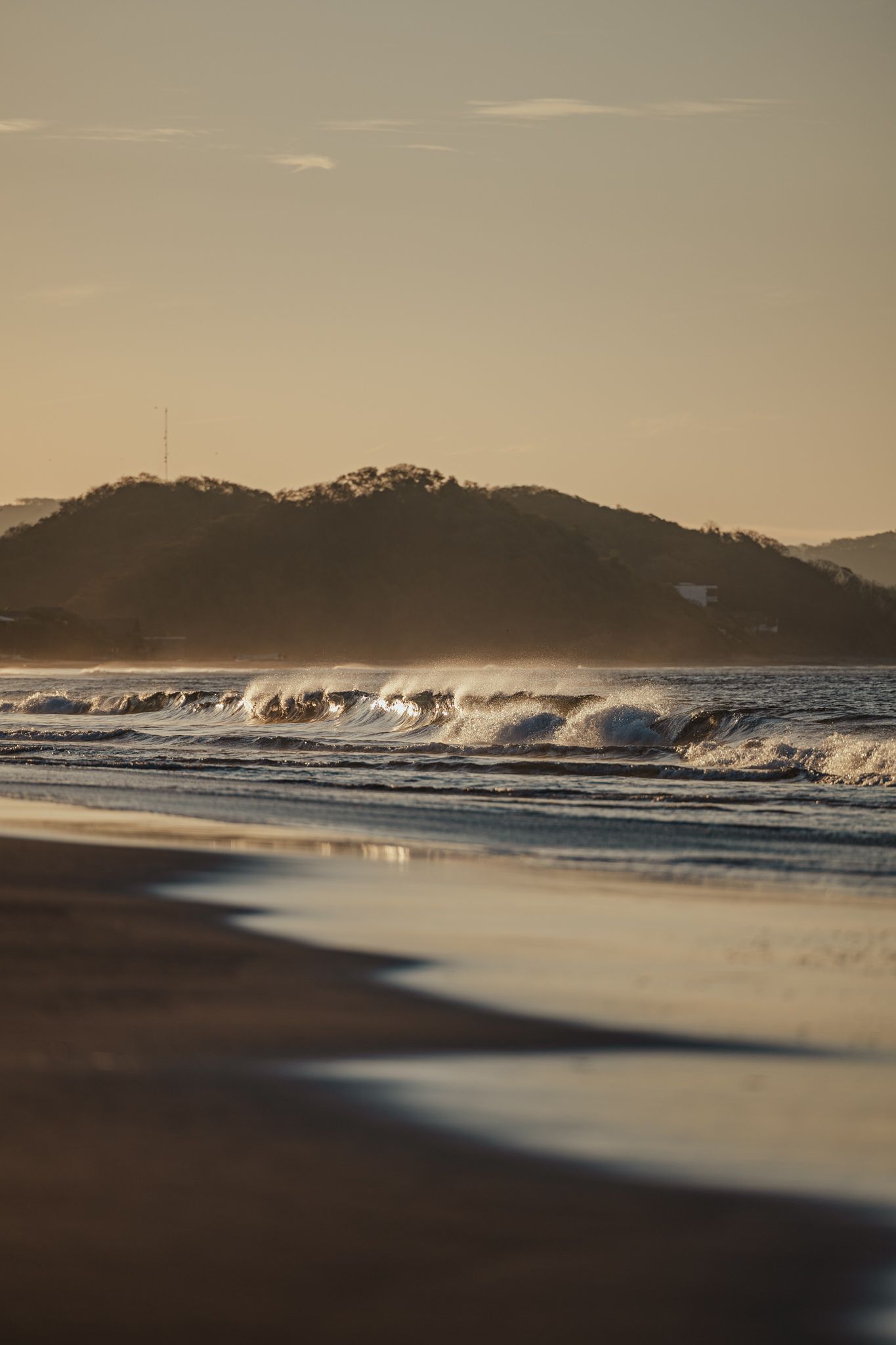 Golden hour waves crash on a sandy beach with misty hills silhouetted against a warm evening sky in the background.