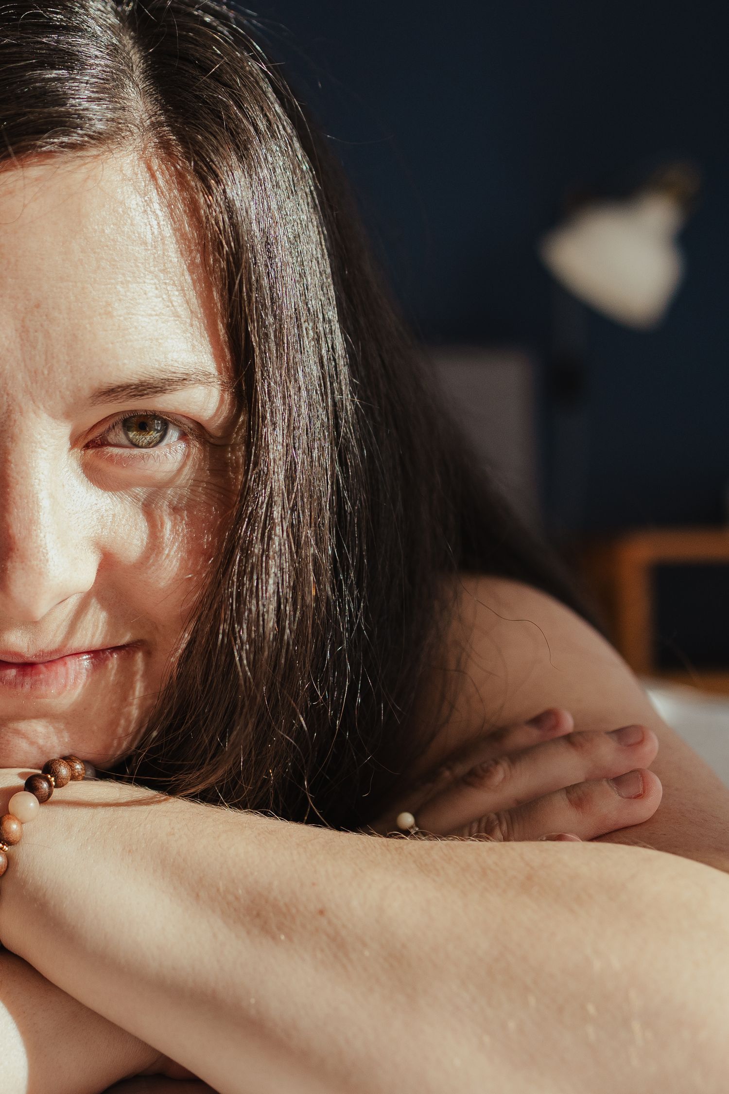 Close-up portrait of a dark-haired woman smiling softly, resting her chin on her arms wearing a beaded bracelet.