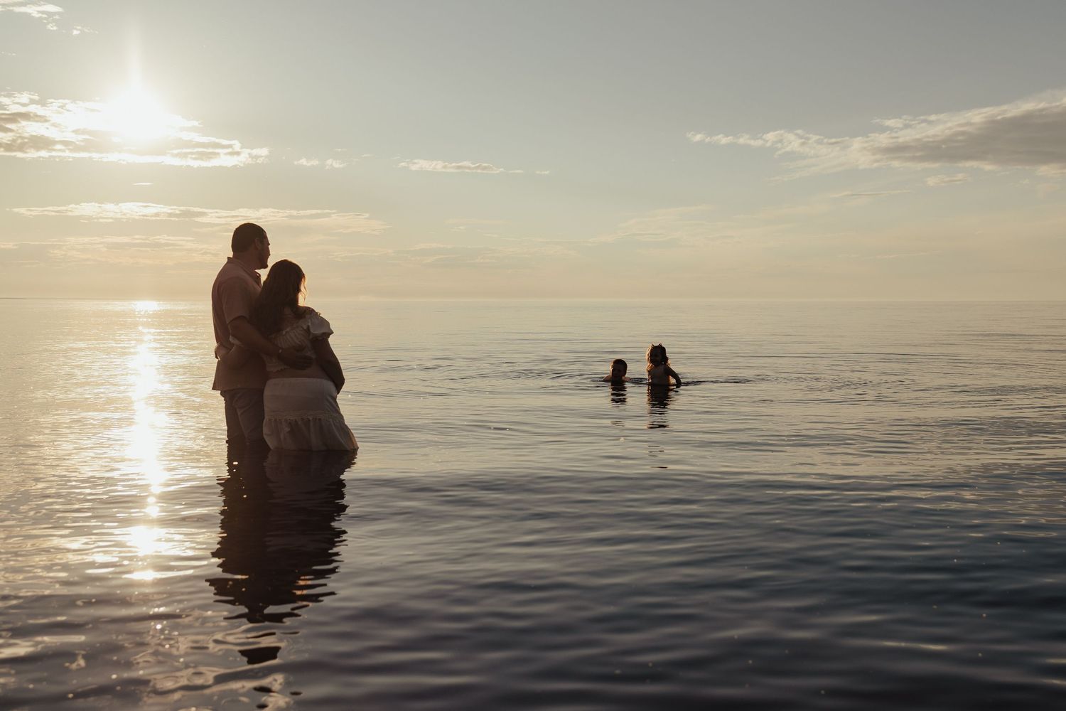Couple standing in calm ocean water at sunset with two people swimming in the background.