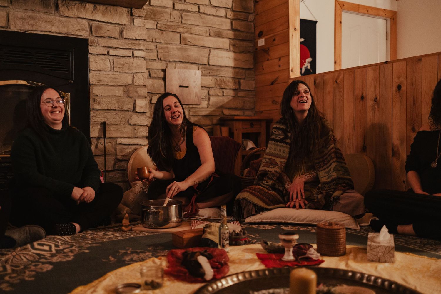 Three women laughing together by a stone fireplace in a cozy rustic cabin setting.