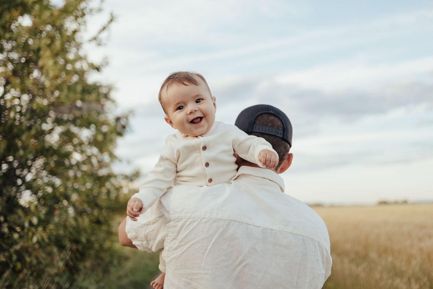 A joyful baby in white outfit being held up by a father in a cap, laughing outdoors in a sunny field.