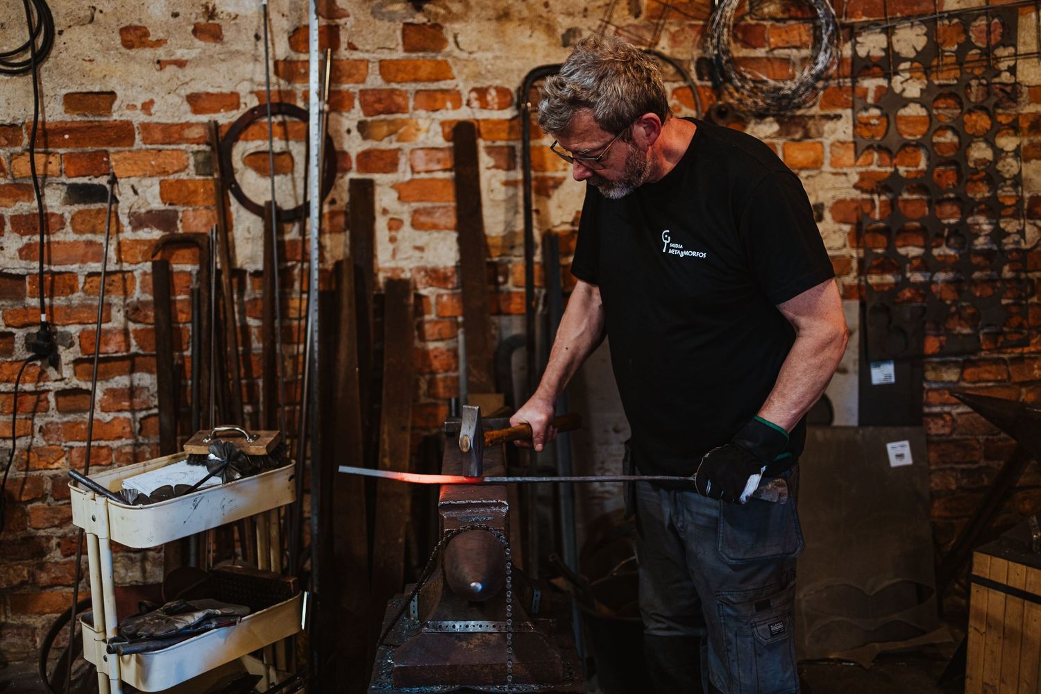 A blacksmith shapes a glowing hot metal blade on an anvil in a rustic brick workshop filled with tools.