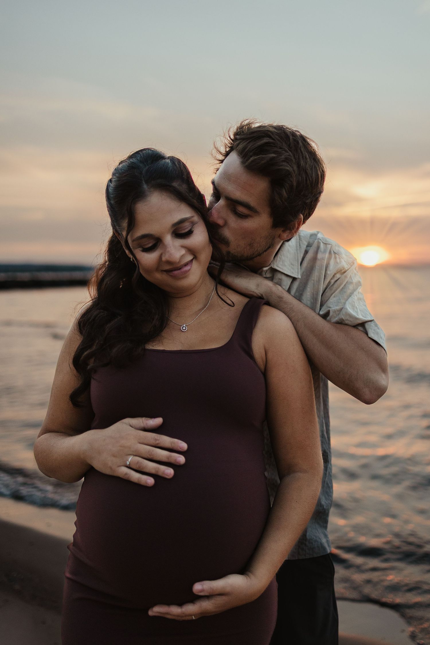Pregnant couple embracing at sunset by the ocean, man kissing woman's temple while they cradle her baby bump.