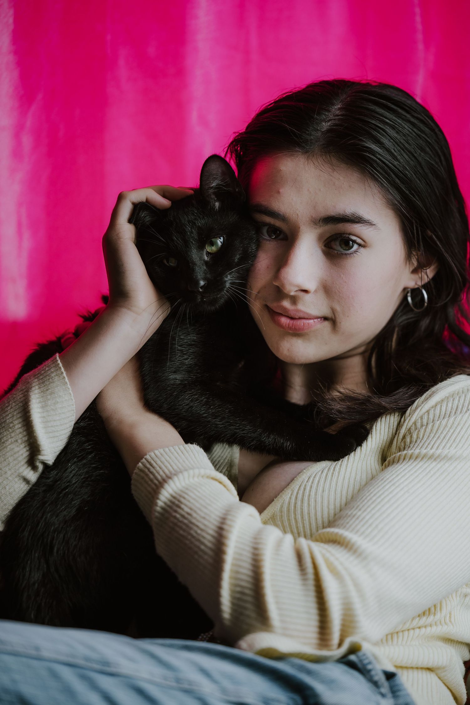 Young woman in cream sweater holding a black cat against a vibrant pink background.