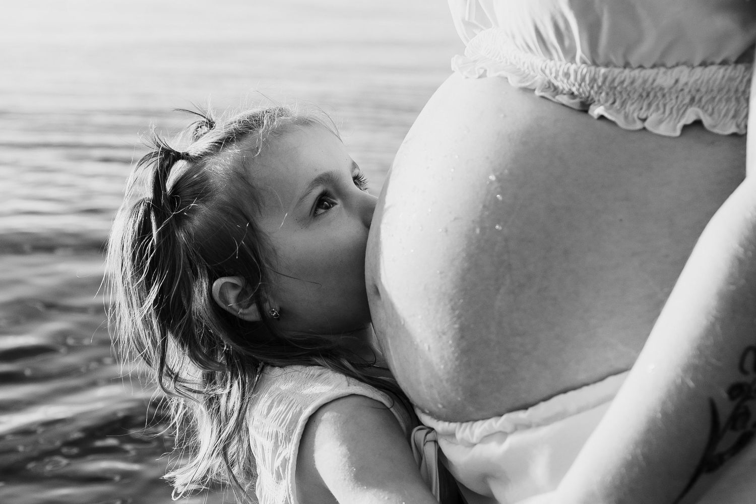 A young girl lovingly kisses a pregnant womans baby bump near a shimmering lake in black and white.