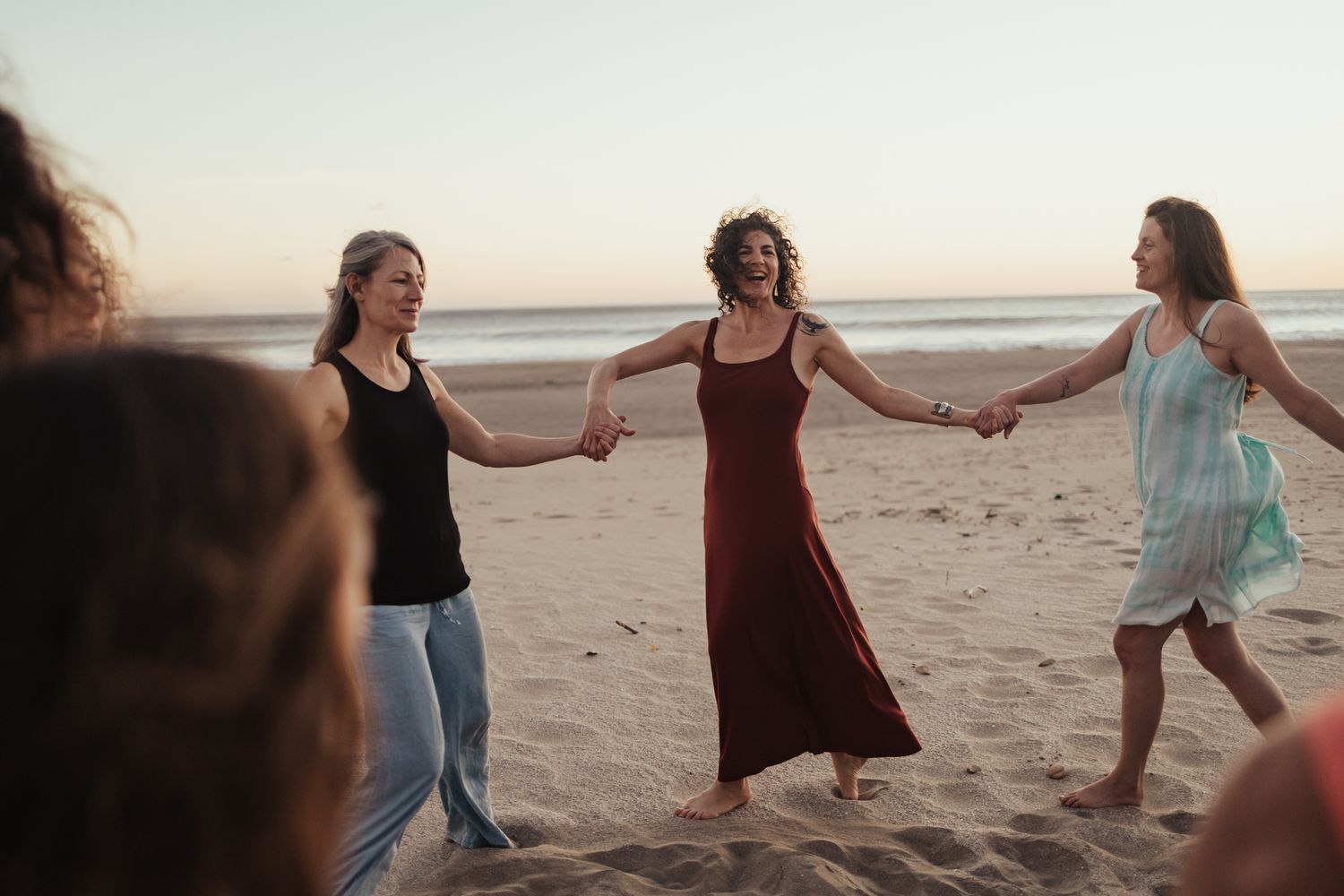 Women holding hands in a circle on a sandy beach at sunset, dancing together in colorful dresses.