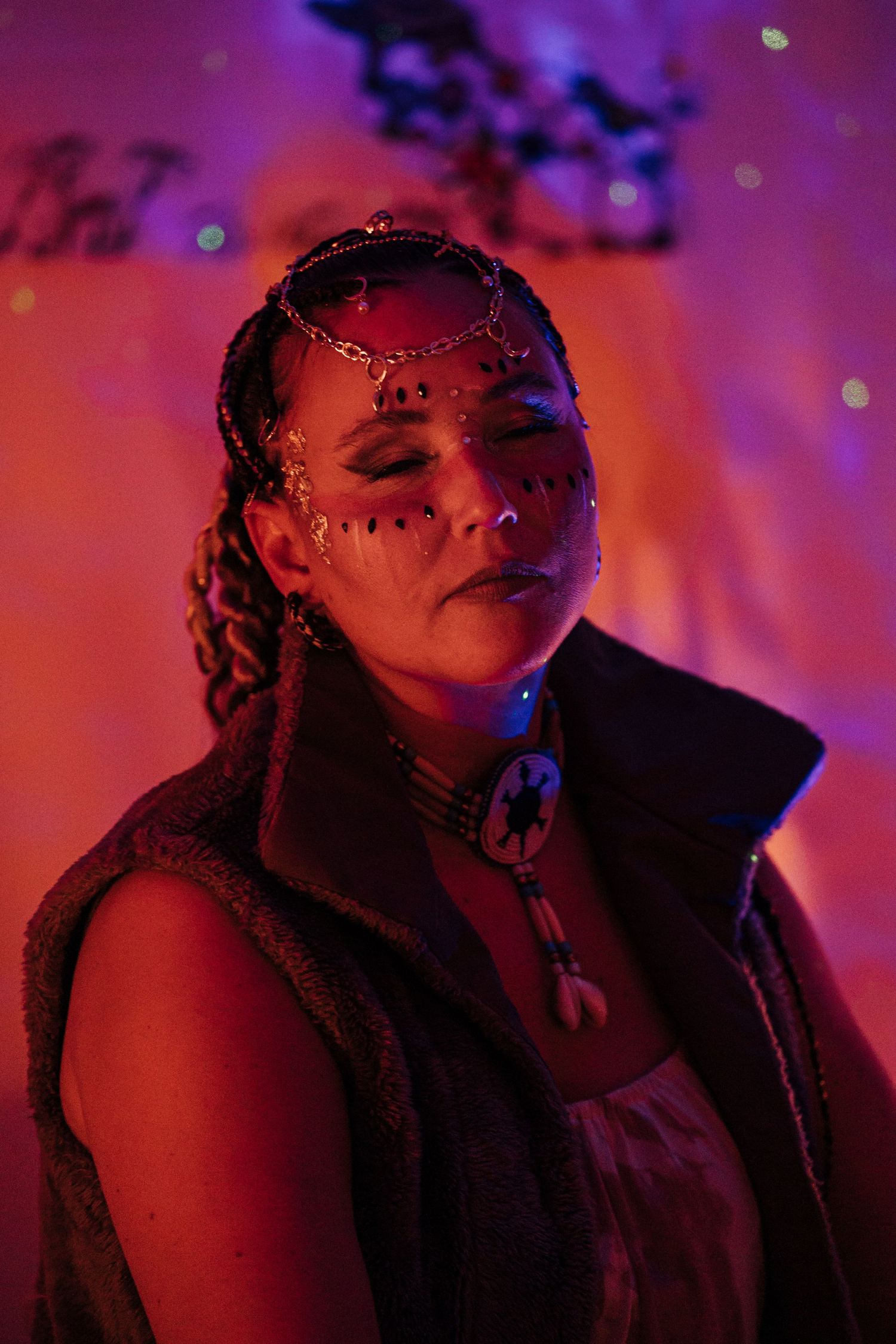 Woman with face gems and crown headpiece poses under purple and red festival lights wearing boho vest and necklace.