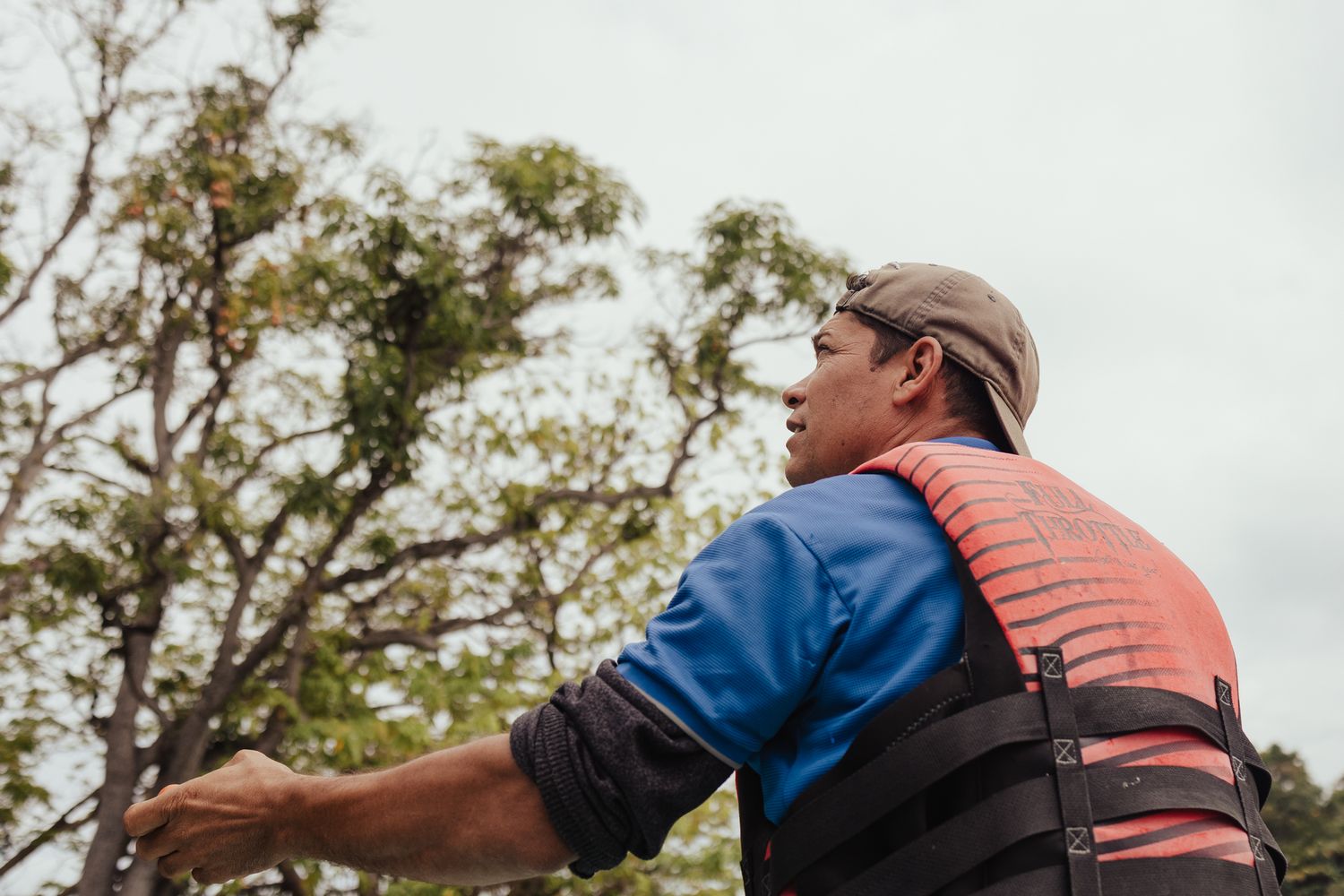 Man in blue shirt and red life vest gesturing outdoors near trees.