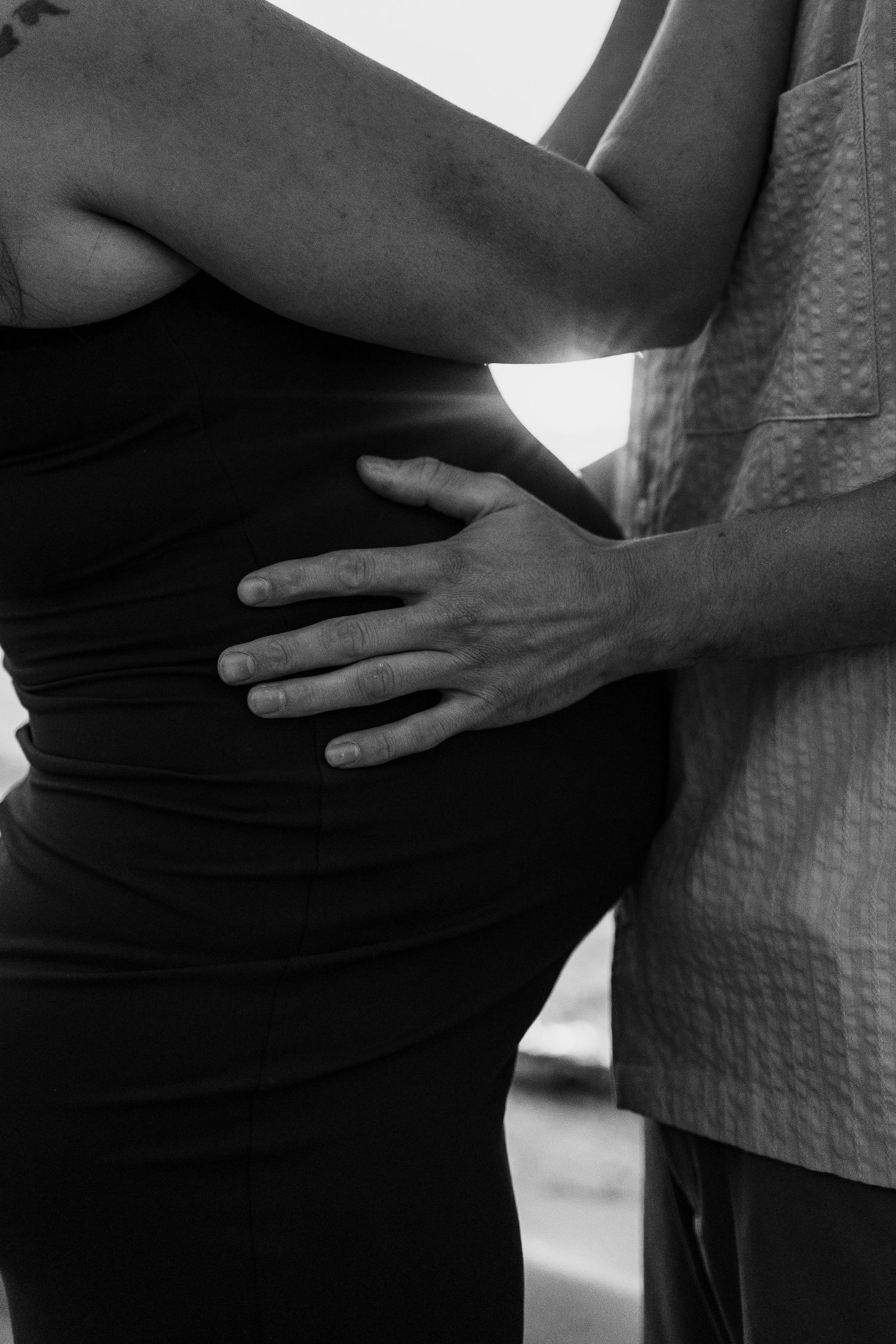 Black and white photo of a partner tenderly holding a pregnant woman's baby bump from behind.