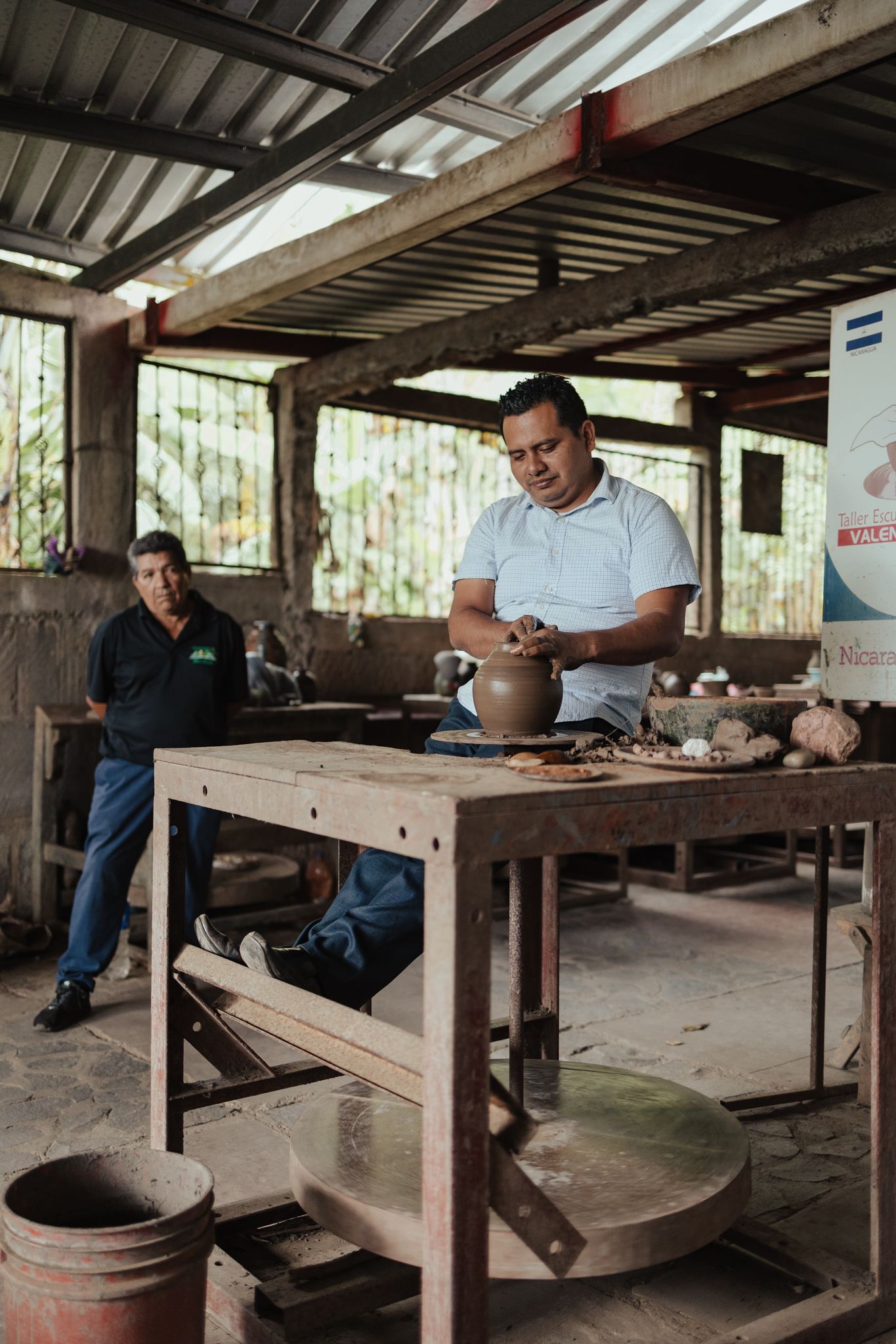 Man shaping clay on a pottery wheel in a rustic open-air workshop with another person watching.