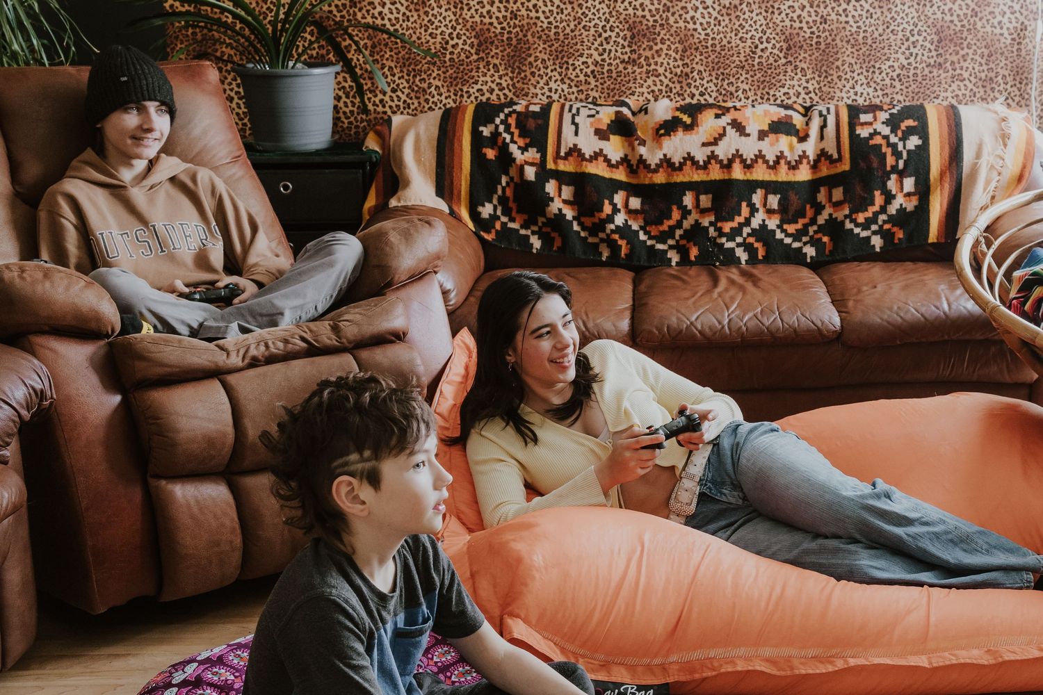 Three people relaxing in a cozy living room with brown leather sofas, playing video games on bean bag chairs.