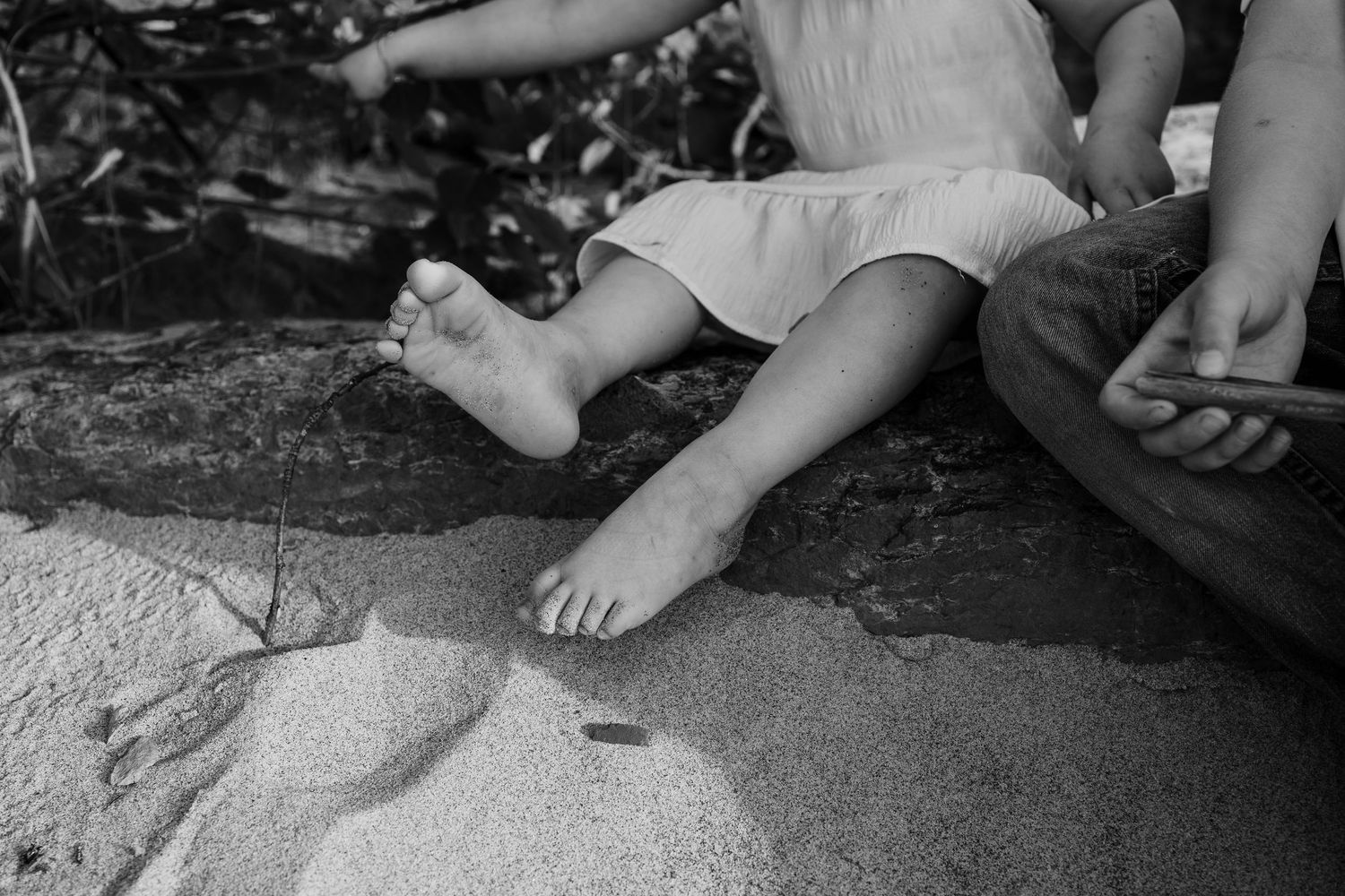 Black and white close-up of two children sitting barefoot on sandy ground near water.