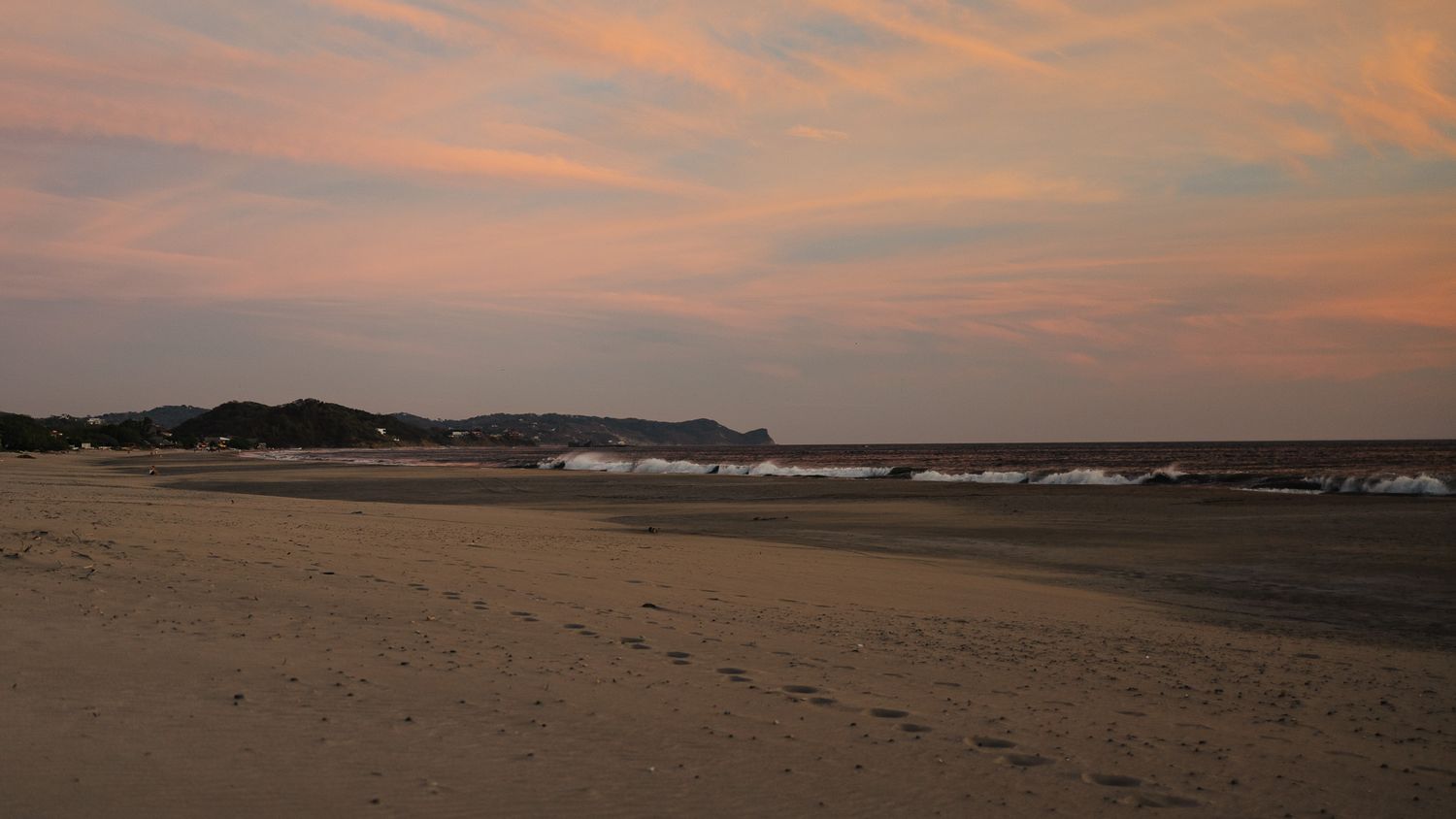 A serene beach at sunset with pink and orange clouds over calm waves and sandy shores with footprints.