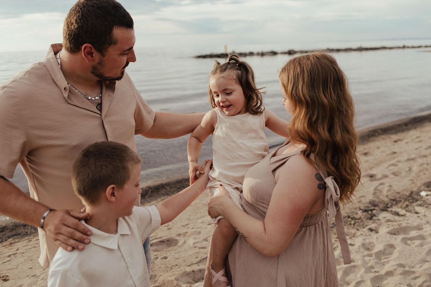 Pregnant woman and man with two young children smiling on a sandy beach during a family maternity photo session.