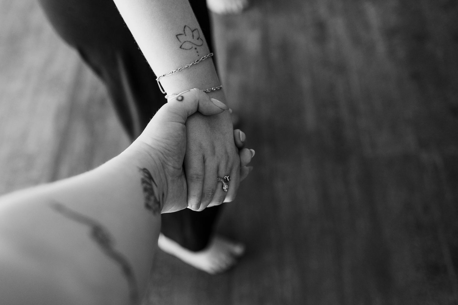 Two people holding hands in black and white, showing tattoos and a bracelet, symbolizing connection and love.