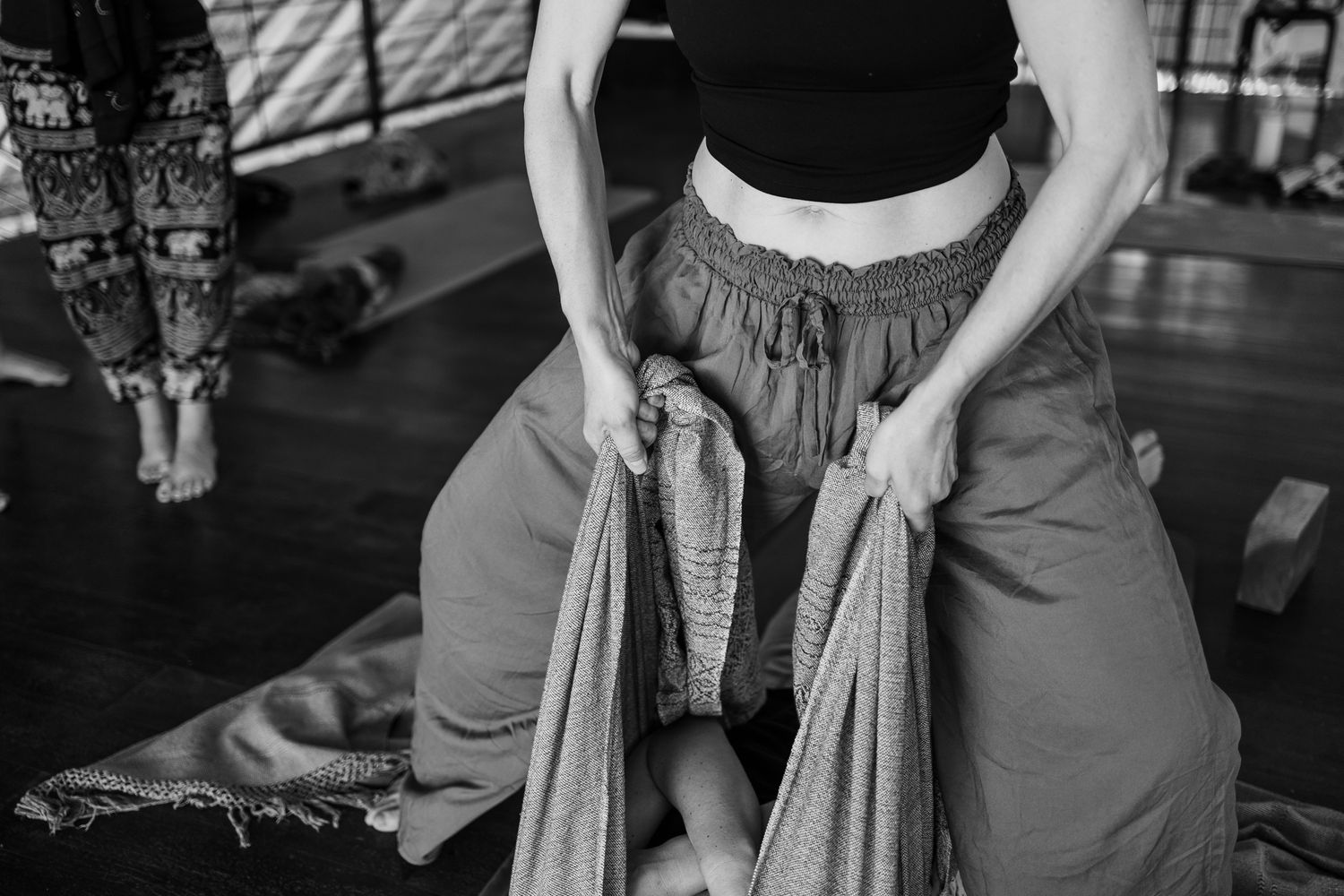 Black and white photo of a woman in loose pants adjusting her clothing during a yoga class.