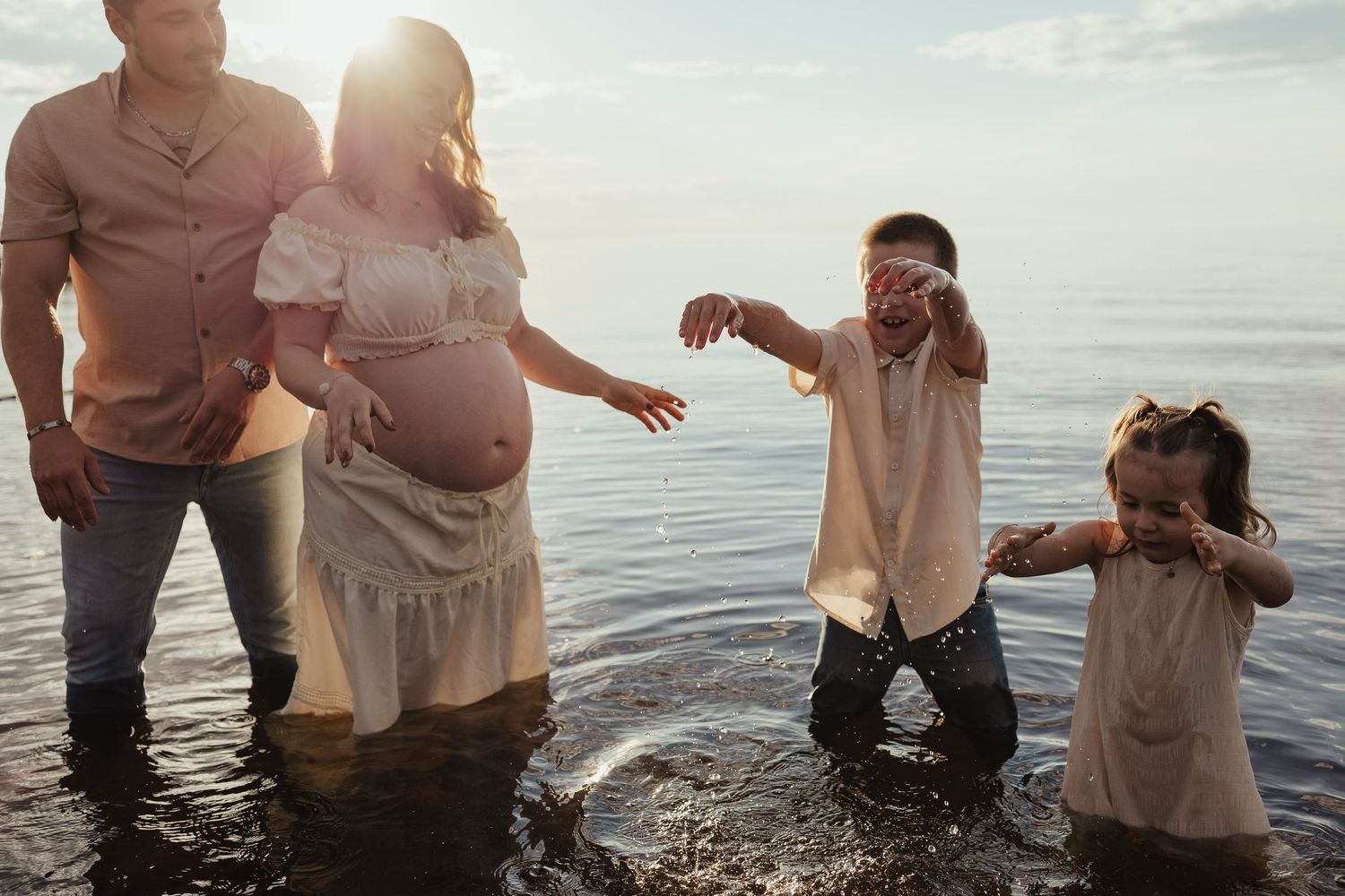 Pregnant mother and father stand in shallow water as their two children play around them.