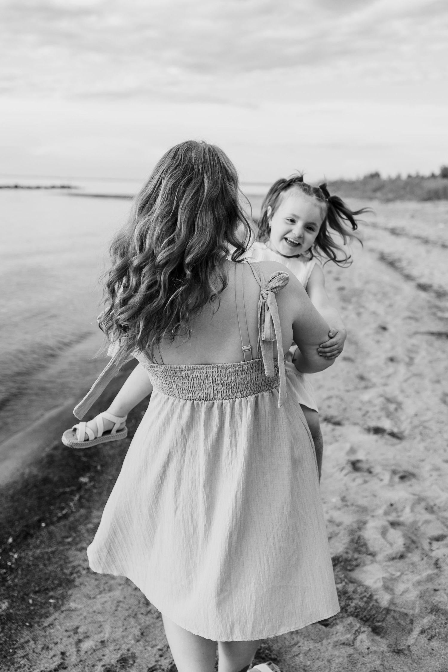 Black and white photo of a mother holding her laughing daughter on a sandy beach near the water.