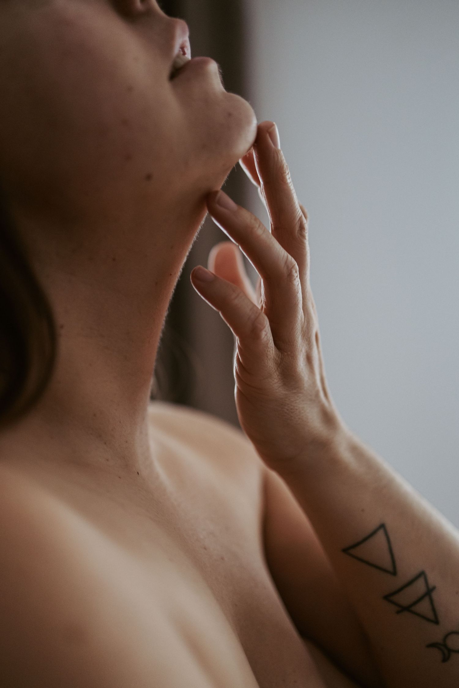 Close-up of a person with geometric triangle tattoo on forearm, hand touching chin against neutral background.