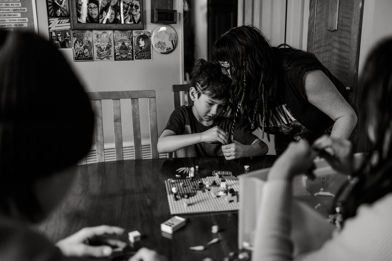 Black and white photo of a child and adult with dreadlocks building with LEGOs at a wooden dining table.