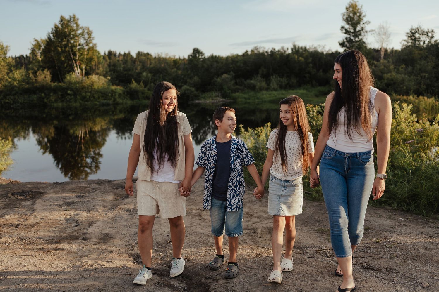 Family of four holding hands and walking near a scenic lake surrounded by lush green trees on a sunny day.