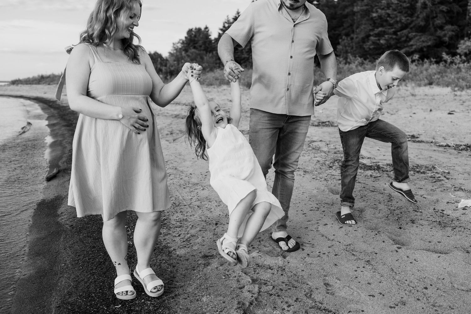 Black and white photo of a family swinging their daughter on a beach while their son plays nearby.
