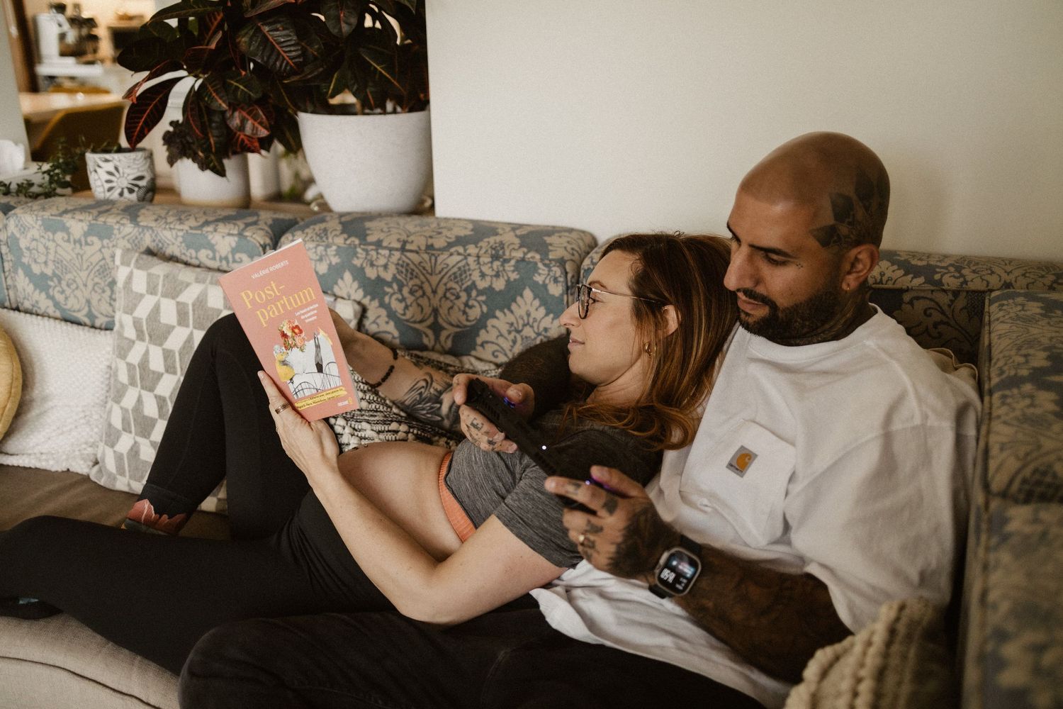 Couple relaxing on couch together, woman lying back reading a book while man holds her lovingly in cozy home setting.