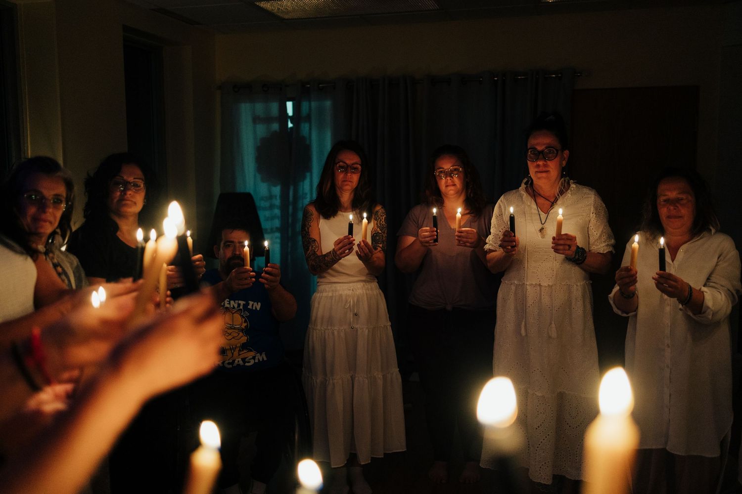 Group of women in white dresses holding candles during a candlelight ceremony in a darkened room.