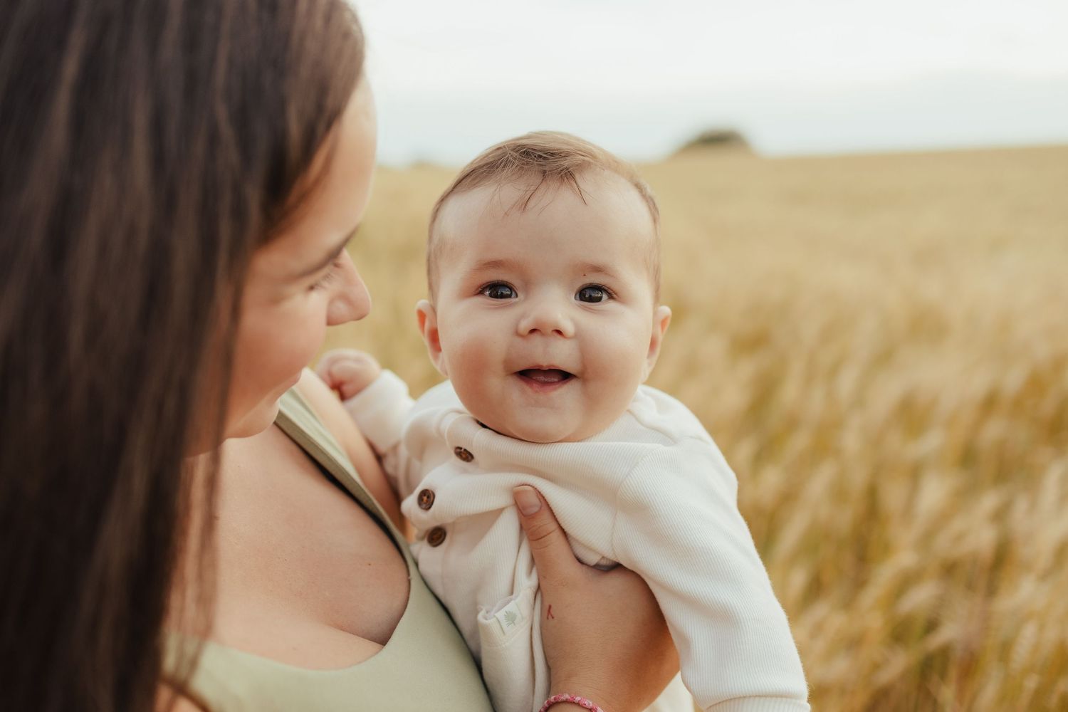 A smiling baby in a white outfit being held by a mother in a golden wheat field on a sunny day.
