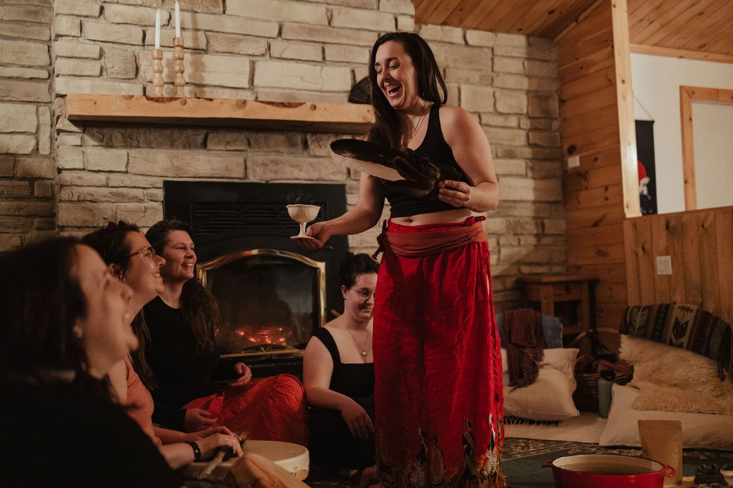 Woman in red skirt laughs while holding a cat near a stone fireplace in a cozy log cabin with friends.