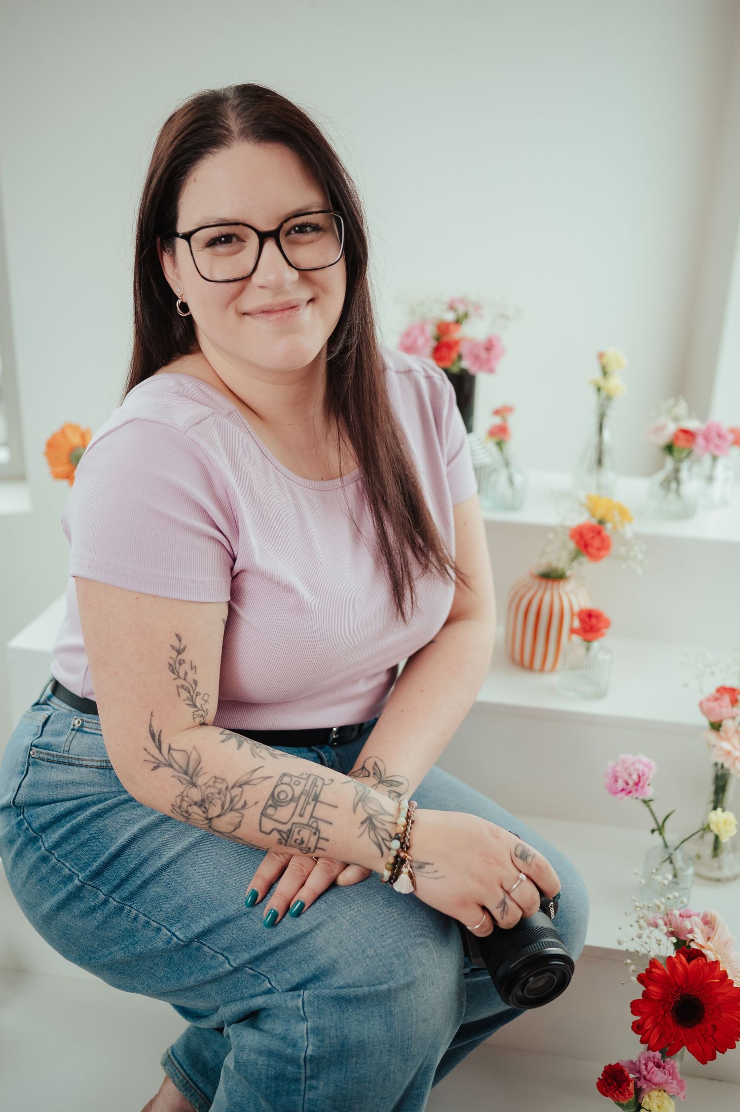 Woman with tattoos and glasses holds camera, sitting among colorful flowers in bright studio setting.